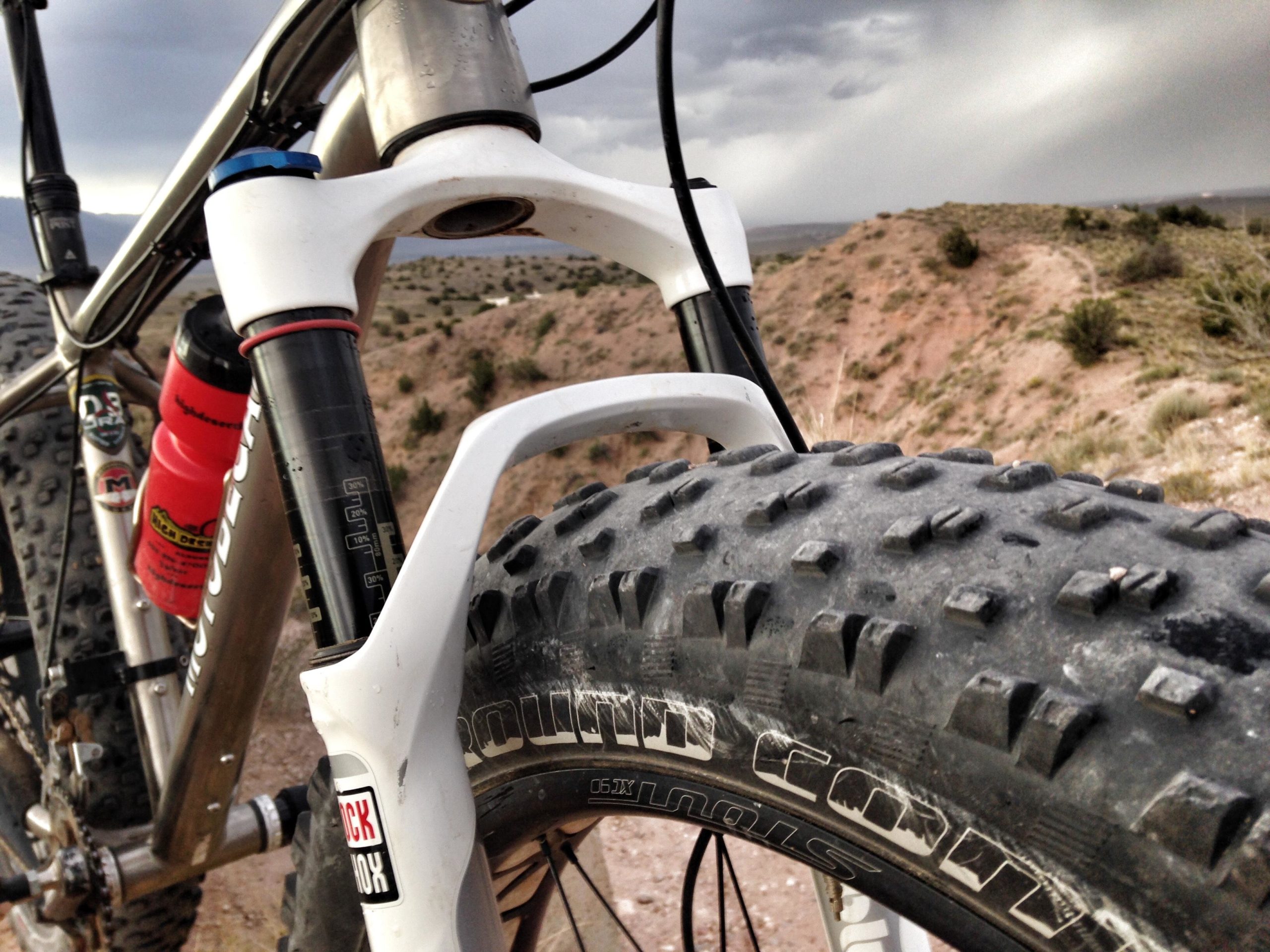 Close-up view of a mountain bike's front fork and tire, showcasing the sturdy design and knobby tread of the tire, set against a rugged outdoor landscape with hills and a cloudy sky in the background. The bike features a metal frame and a visible water bottle attached to it. Mariposa Fat Bike Trails mountain bike trail.