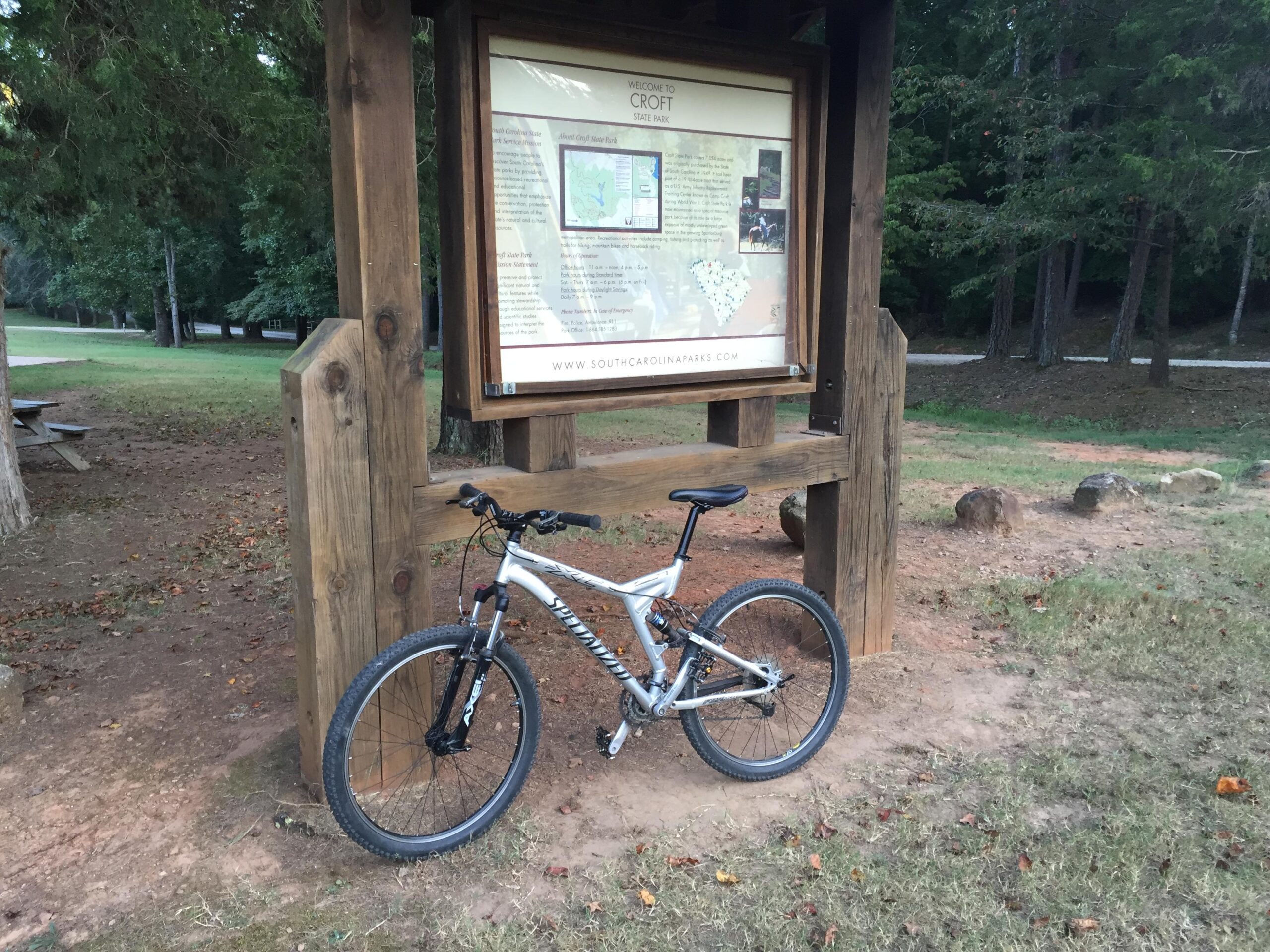Specialized FSRxc Pro: A mountain bike leaning against a wooden park information sign in a grassy area surrounded by trees. The sign includes a map and details about Croft State Park. There are a few rocks visible on the ground, and a road can be seen in the background.