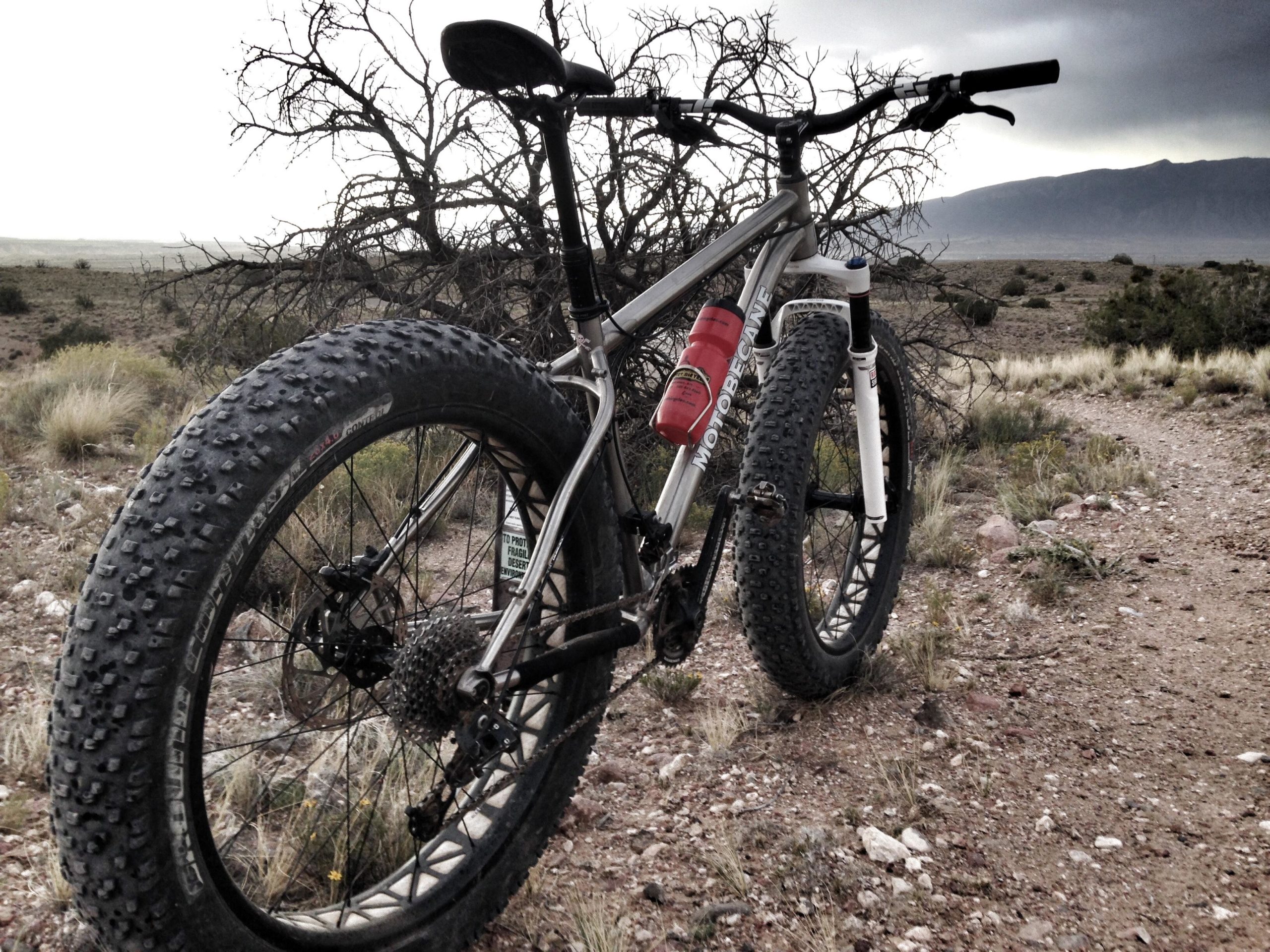 A close-up view of a fat bike resting on a rocky trail, with a desert landscape in the background. The bike features thick, knobby tires and a water bottle attached to the frame, while distant mountains and a cloudy sky are visible in the horizon. Mariposa Fat Bike Trails mountain bike trail.