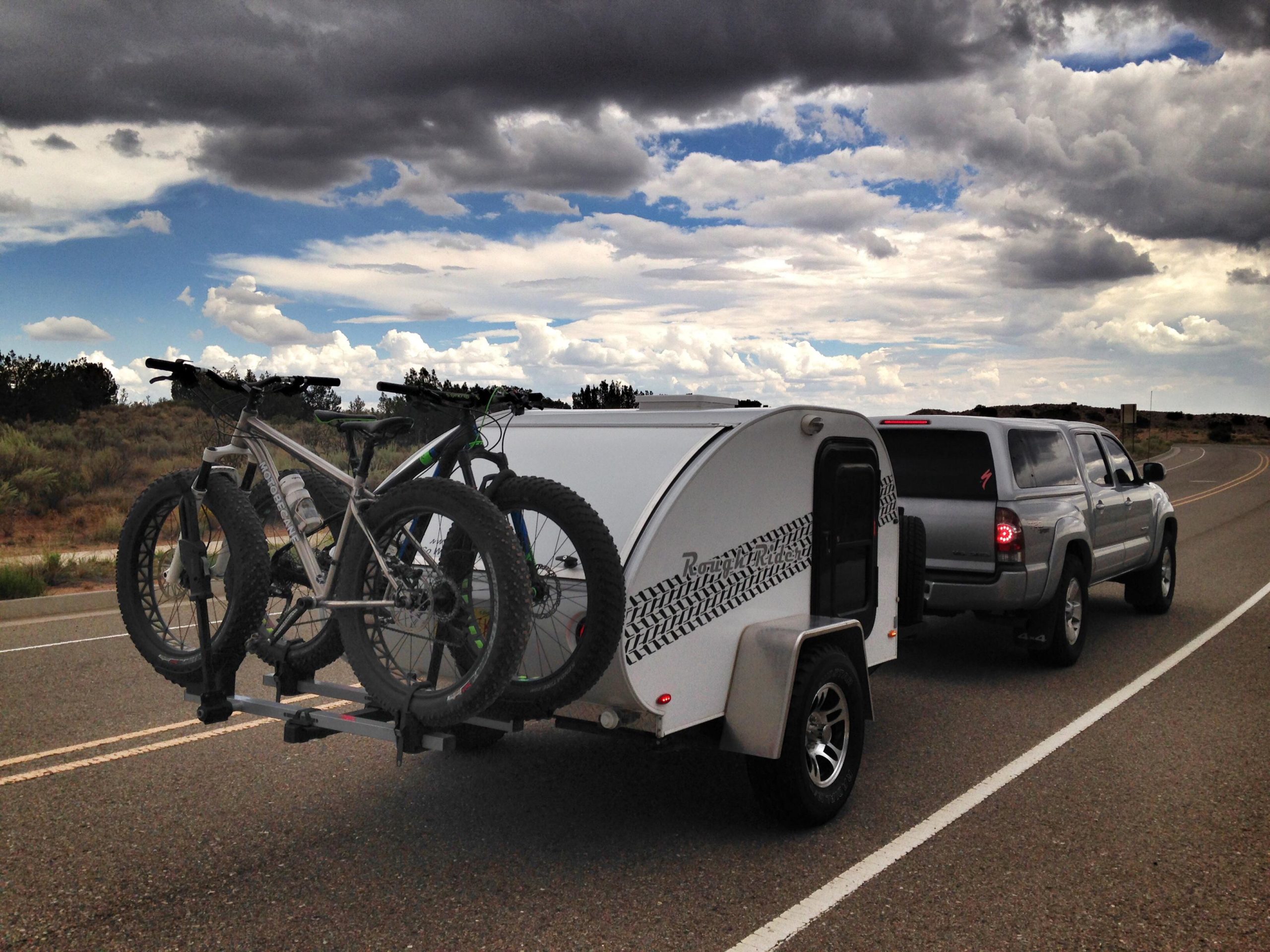 A silver pickup truck towing a small white trailer with a tire tread design, carrying two mountain bikes on a bike rack. The scene features a wide, open road with a cloudy sky, showcasing an expansive landscape with shrubs and trees in the background. Mariposa Fat Bike Trails mountain bike trail.