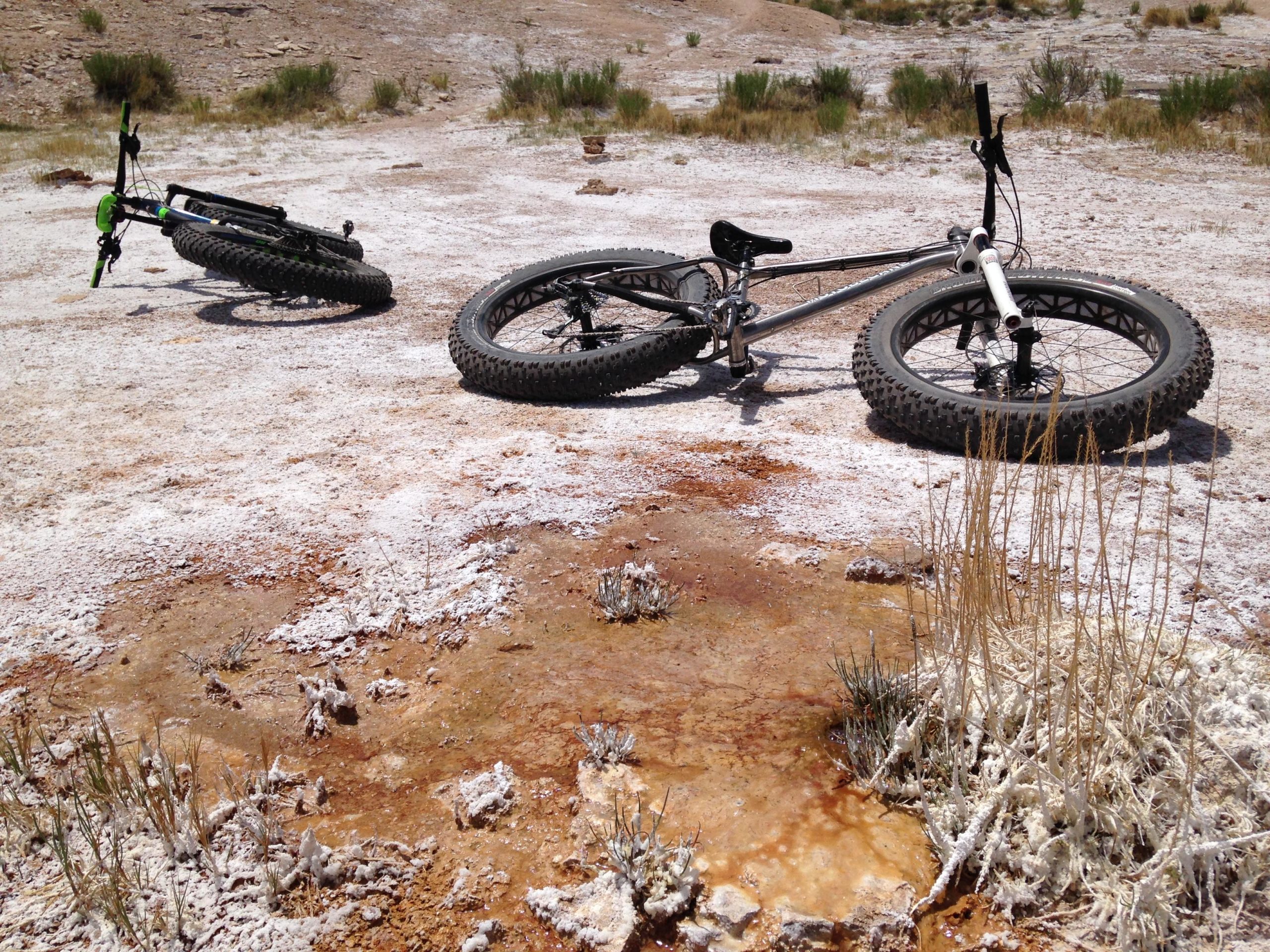 Two fat-tire bicycles lie on a dry, rocky surface with patches of white salt and reddish-brown soil. Sparse vegetation, including small grass and plants, is visible in the background, indicating a rugged outdoor terrain. The scene captures a sunny day, showcasing a desolate yet striking landscape. White Ridge Bike Trails mountain bike trail.