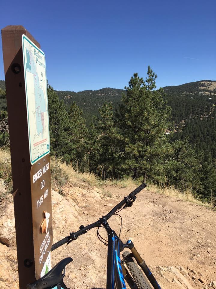 A bicycle leaning against a post with a trail sign, indicating that bikes must go a certain way. In the background, a scenic view of lush green mountains and a clear blue sky. Betasso Preserve mountain bike trail.
