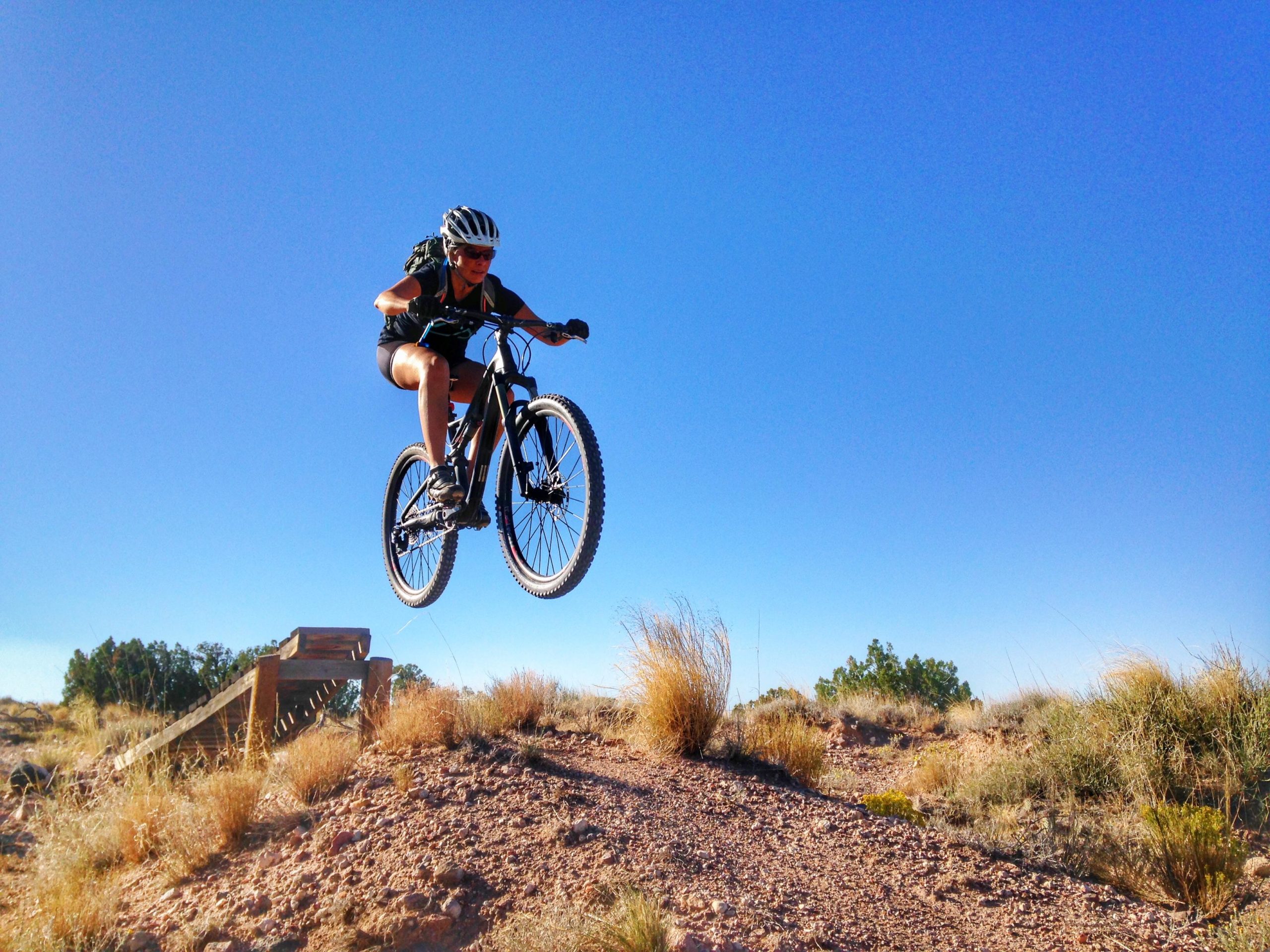 A person wearing a helmet and athletic gear is mid-air, jumping off a wooden ramp on a mountain bike. The scene is set against a clear blue sky, with dry desert terrain and sparse vegetation in the background. Mariposa Fat Bike Trails mountain bike trail.