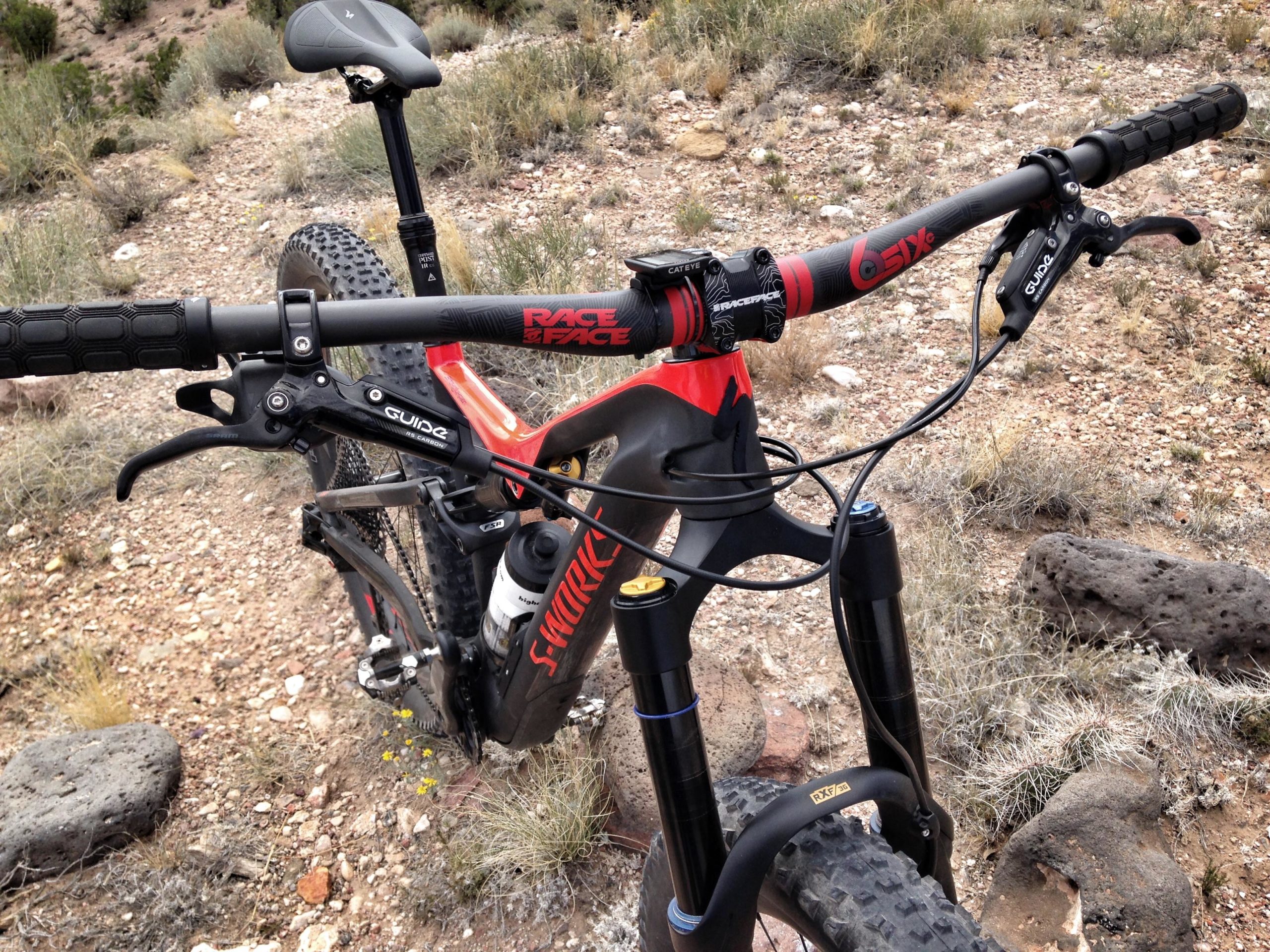 Close-up view of a mountain bike's handlebars, showcasing the grips, brake levers, and suspension fork. The bike is positioned on a natural dirt trail with rocks and sparse vegetation in the background, indicating an outdoor setting suitable for biking. Mariposa Fat Bike Trails mountain bike trail.