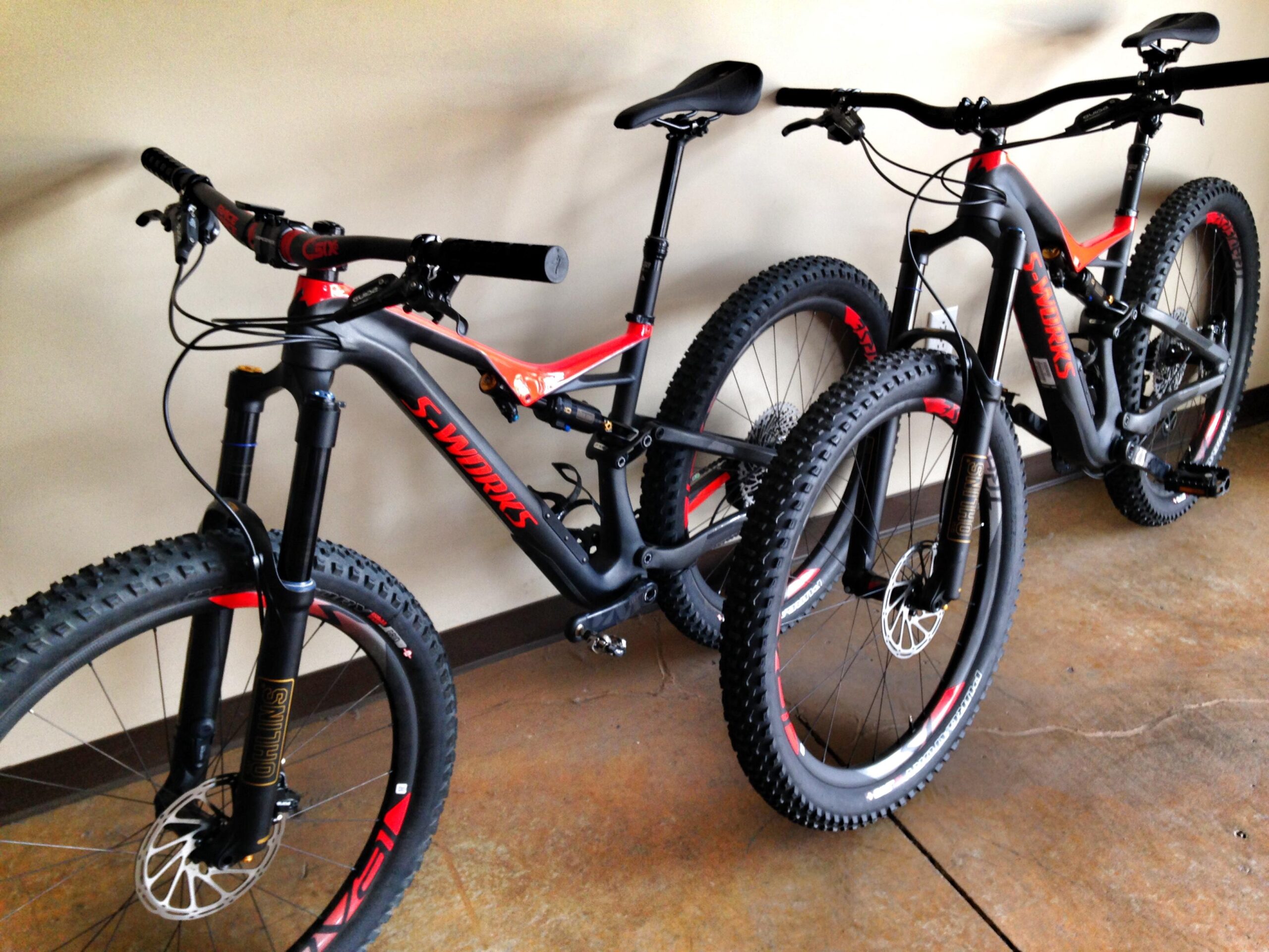 Two high-end mountain bikes are leaning against a wall. Both bikes feature black frames with red accents, fat tires, and rugged wheels designed for off-road terrain. The handlebars are equipped with various controls, and the bikes are positioned in a well-lit indoor space with a polished concrete floor.