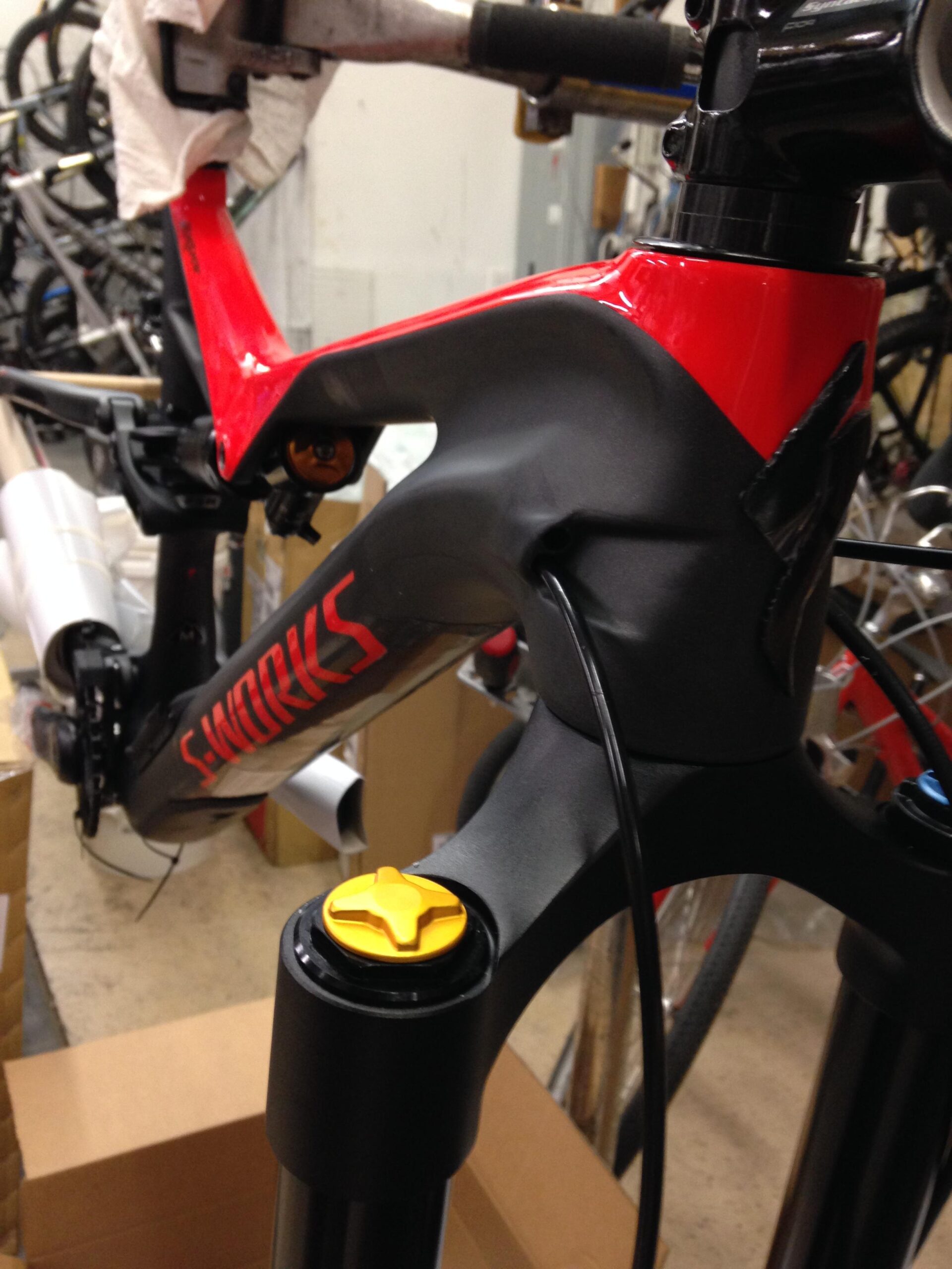 Close-up view of a specialized bike frame in a workshop setting, showcasing a sleek black and red design with a prominent yellow adjustment knob on the front fork. Various bicycles and cardboard boxes can be seen in the background, indicating a busy repair or assembly area.