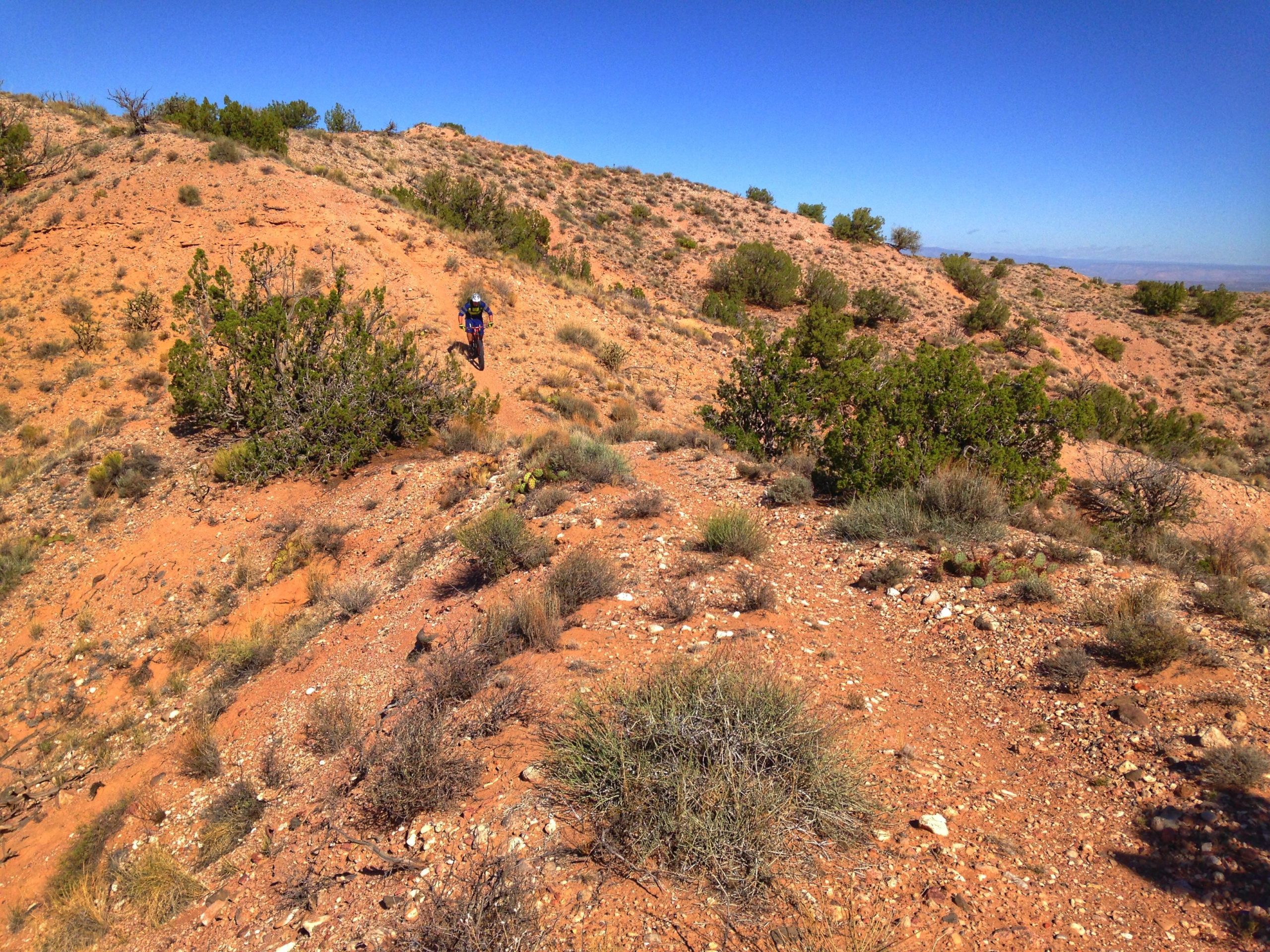 A mountain biker rides along a dusty trail on a rugged landscape characterized by dry, reddish soil, scattered rocks, and sparse vegetation under a clear blue sky. Mariposa Fat Bike Trails mountain bike trail.