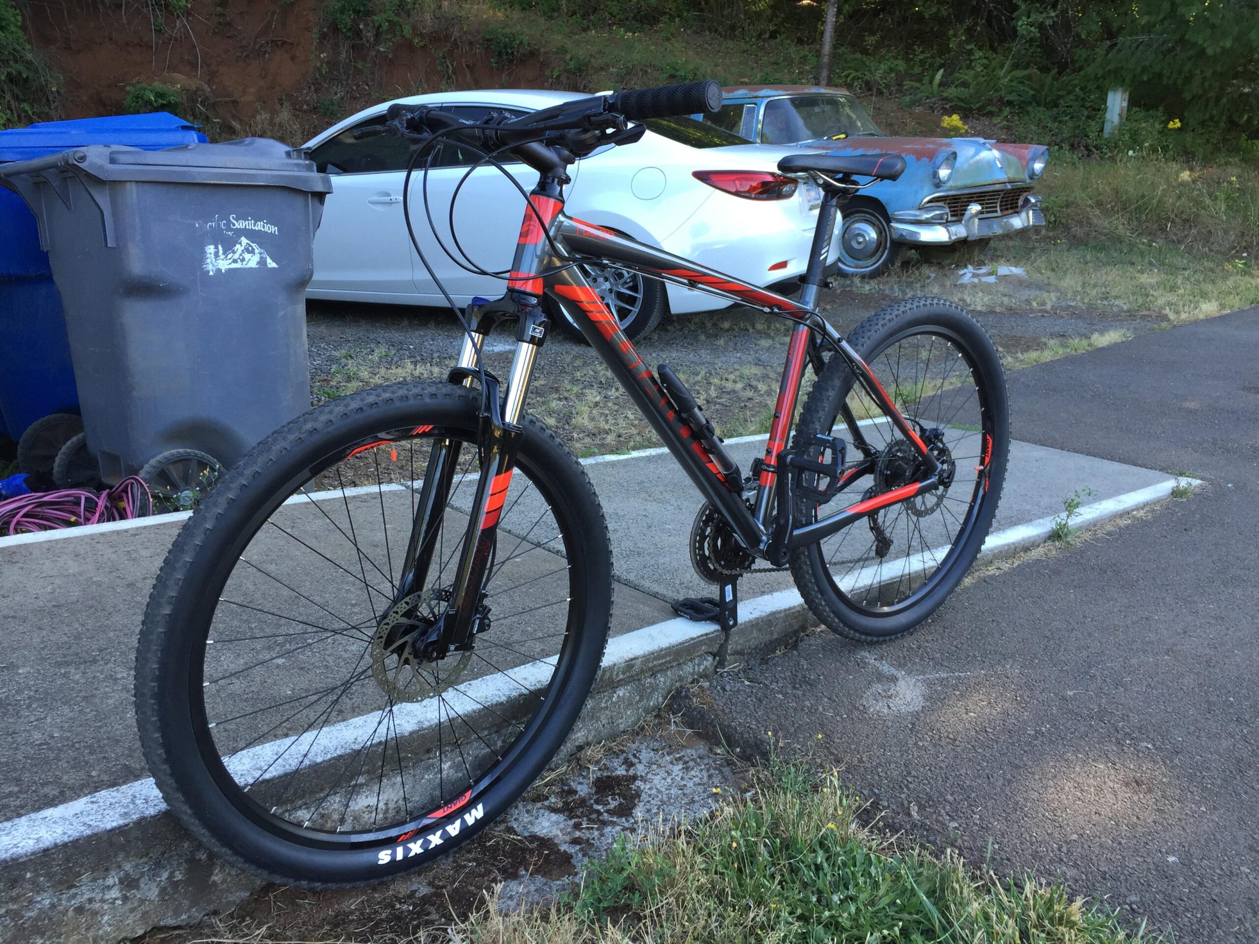 Giant Talon 27.5 3: A mountain bike with a black and orange frame parked on a sidewalk, next to a gray trash bin. In the background, there is a white car and a vintage blue and rusted car, surrounded by grass and trees.