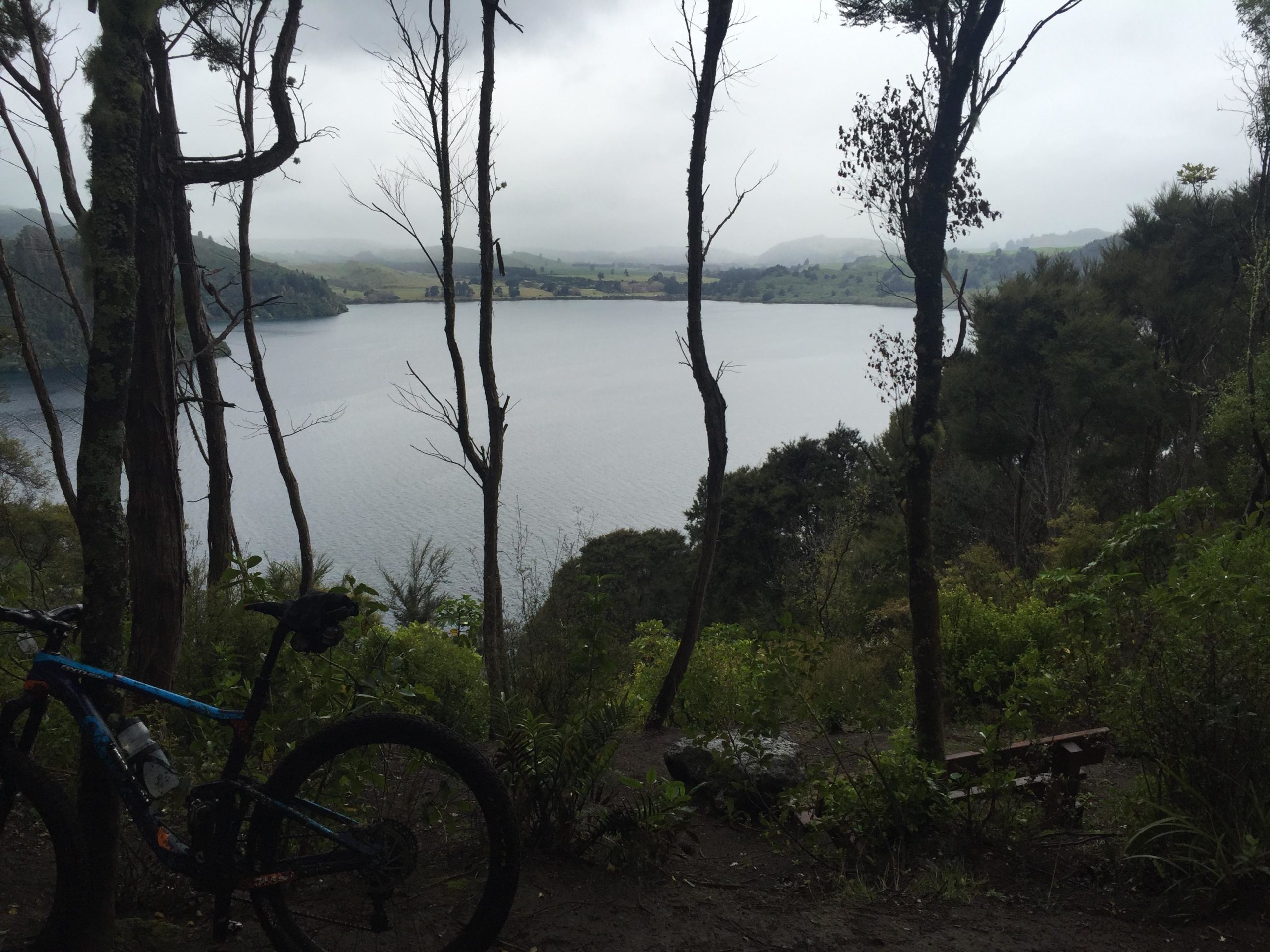 A scenic view of a lake surrounded by lush greenery and trees, with a mountain bike in the foreground. The sky is overcast, creating a tranquil atmosphere. The lake reflects the muted colors of the landscape, and a wooden bench is partially visible to the right, inviting relaxation in nature. Great Lake Trails mountain bike trail.