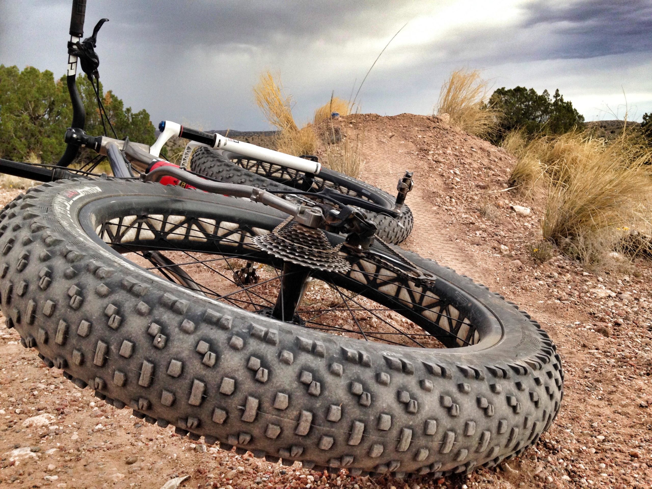 Close-up view of a fat tire bicycle resting on a dirt trail, with the bike's handlebars and gear visible. The background features a partly cloudy sky and sparse vegetation on the trail, indicating a rugged outdoor setting. Mariposa Fat Bike Trails mountain bike trail.