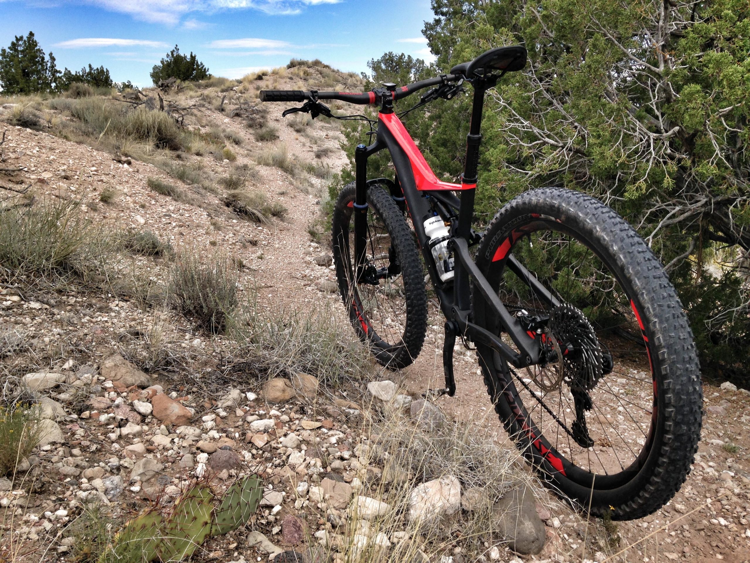 A mountain bike with a black and red frame positioned on a rocky dirt trail surrounded by sparse vegetation and trees under a blue sky. Mariposa Fat Bike Trails mountain bike trail.
