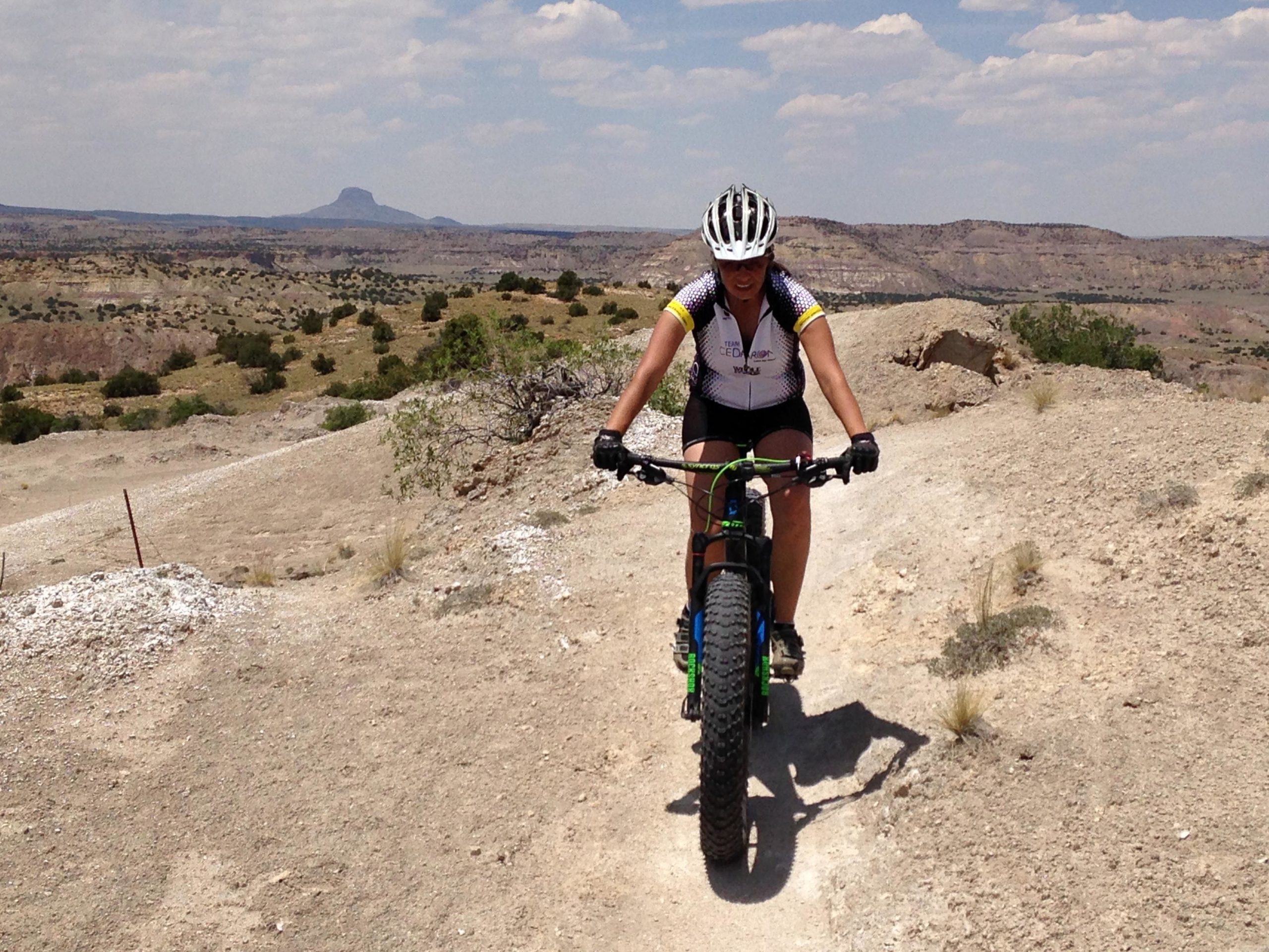 A female mountain biker riding on a dirt trail with a rocky landscape in the background, featuring mountains and sparse vegetation under a blue sky with scattered clouds. The biker is wearing a helmet and a cycling jersey, focused on the trail ahead. White Ridge Bike Trails mountain bike trail.