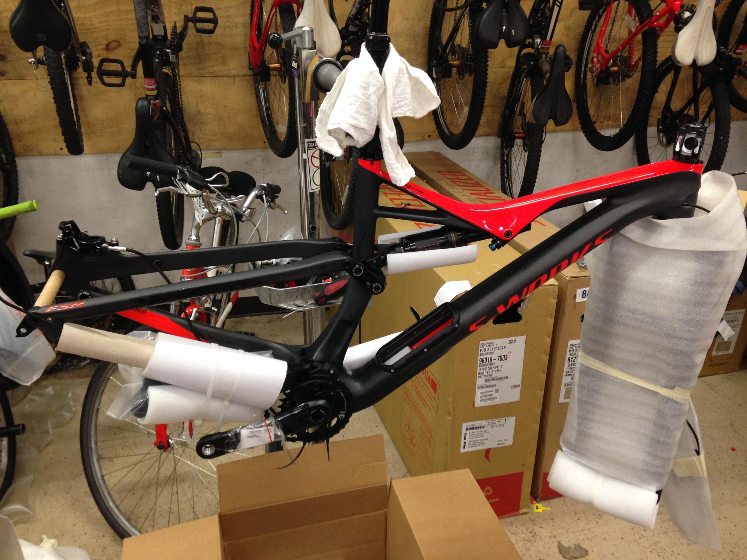 A partially assembled bicycle frame is displayed in a workshop setting, surrounded by packaging materials and boxes. The bike features a sleek black and red design, with protective wrapping on various components. Several bicycles can be seen hanging on a wall in the background.