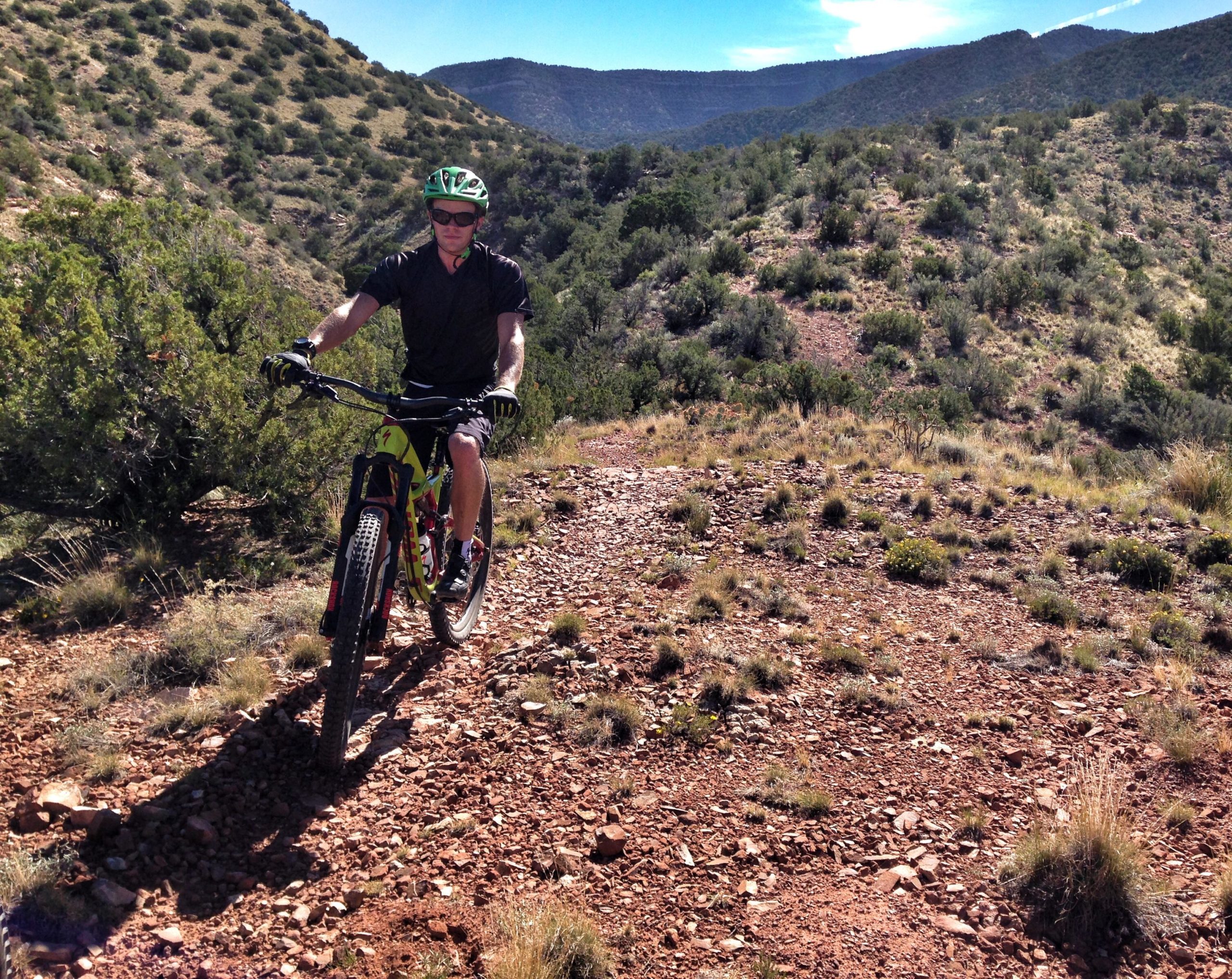 A person riding a mountain bike on a rocky trail surrounded by sparse vegetation and rolling hills under a clear blue sky. The cyclist is wearing a green helmet and sunglasses, dressed in a black shirt and shorts, and is actively navigating the rugged terrain. Placitas Trails mountain bike trail.