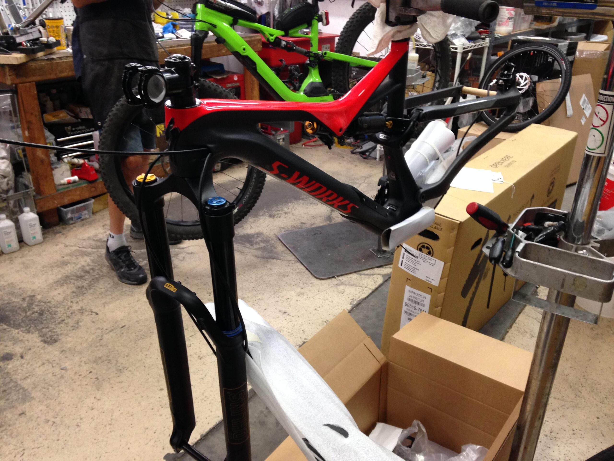 A close-up view of a mountain bike in a workshop, featuring a red and black S-Works frame and fork. The image shows various bike components and tools in the background, with a partially visible green bike also present. Cardboard boxes and other cycling accessories are scattered around the workspace.