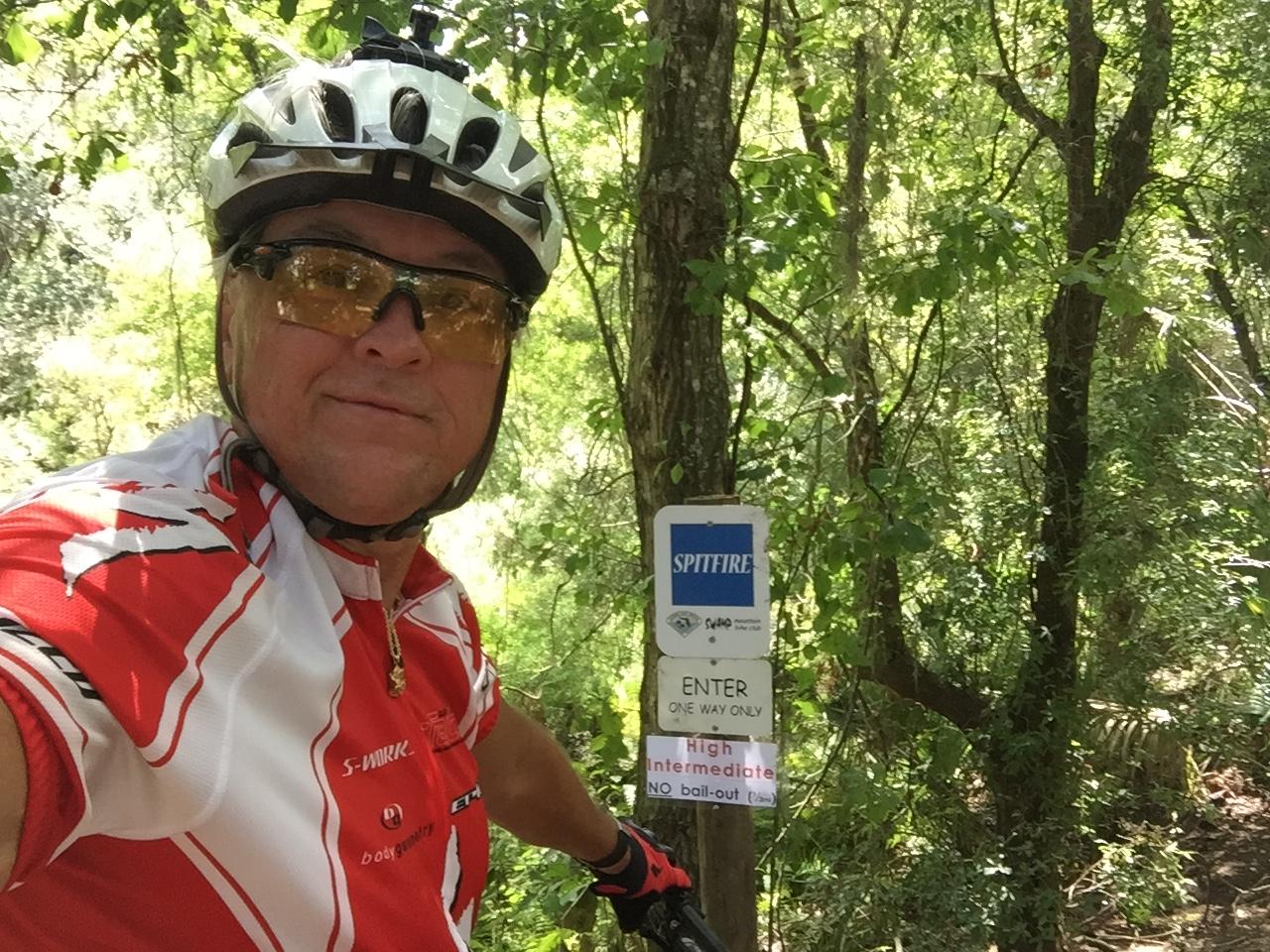 A mountain biker in a red and white cycling jersey and helmet poses for a selfie in a wooded area. A trail sign reads "SPITFIRE" with instructions for entering the trail, indicating it is for high intermediate riders only. The surrounding greenery suggests a sunny day in nature. Alafia River State Park mountain bike trail.