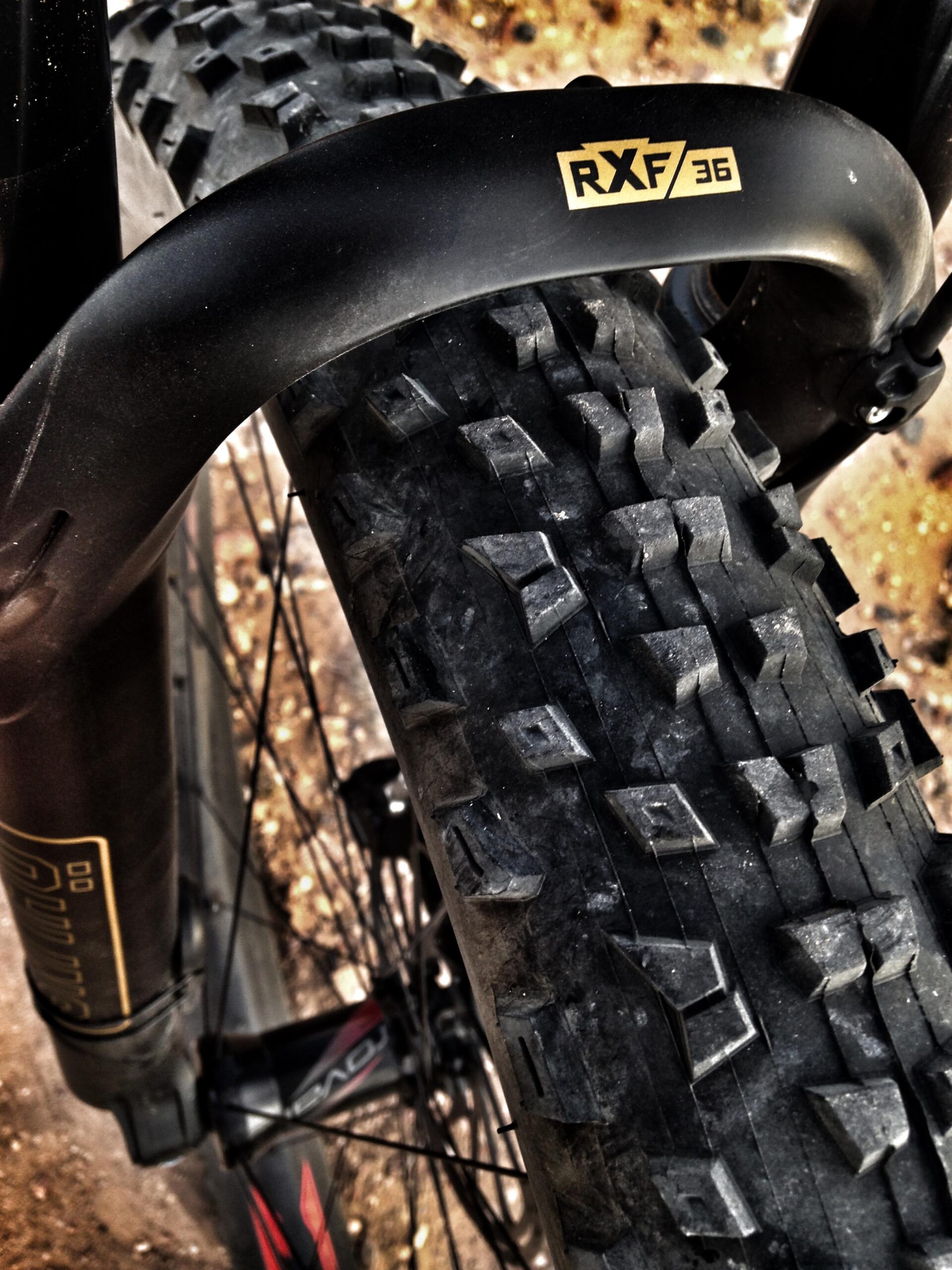 Specialized Stumpjumper FSR 6Fattie: Close-up view of a mountain bike's front tire and suspension fork. The tire features a rugged tread pattern designed for off-road performance, while the fork is labeled "RXF 36." The image captures the details of the tire's texture and the sleek design of the fork against a blurred background of dirt and gravel.