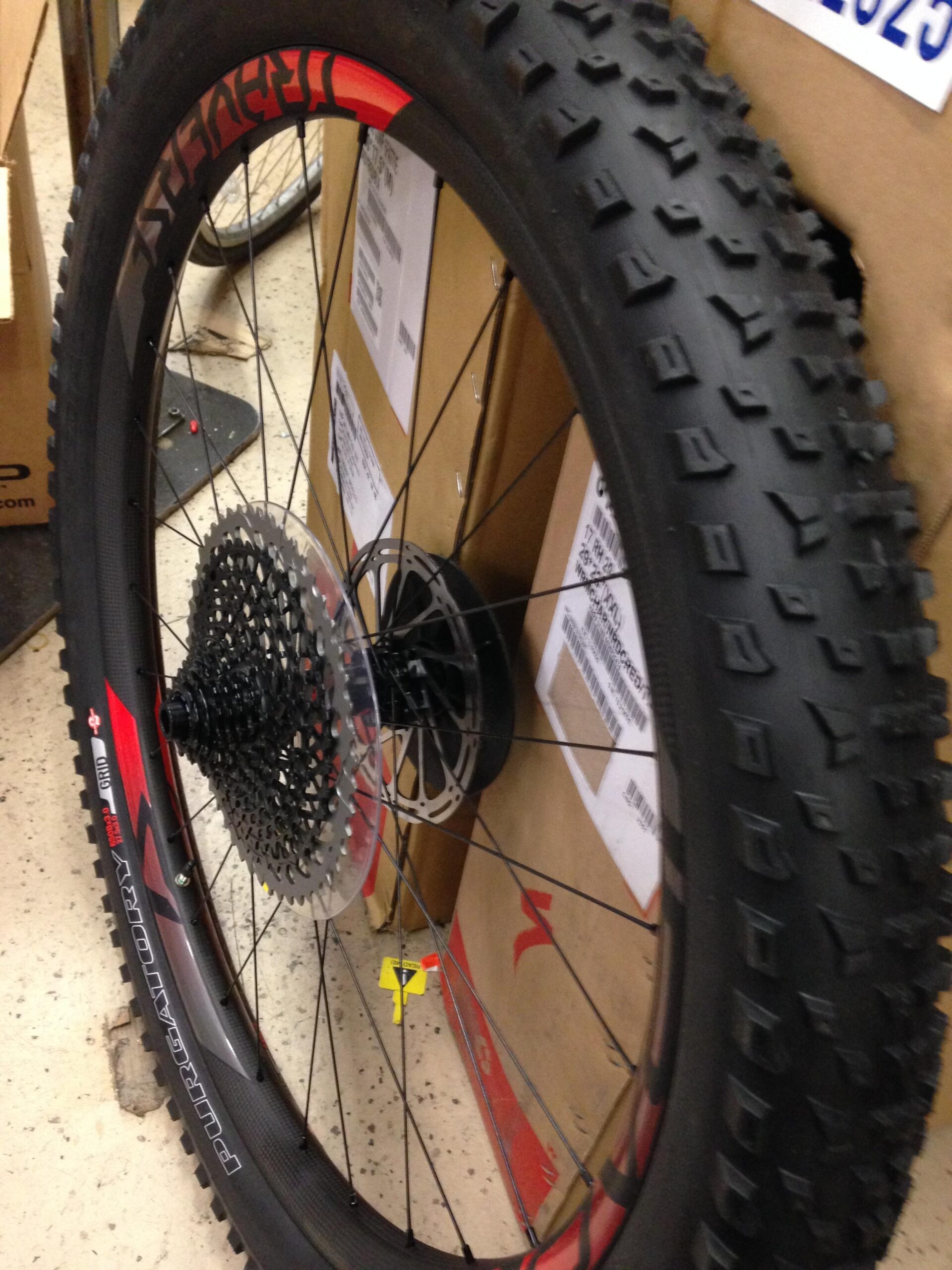 Close-up view of a bicycle wheel with a thick, knobby tire mounted on a black rim. The wheel features a cassette gear system and is positioned against a backdrop of cardboard boxes. The tire has distinctive tread patterns designed for off-road use.