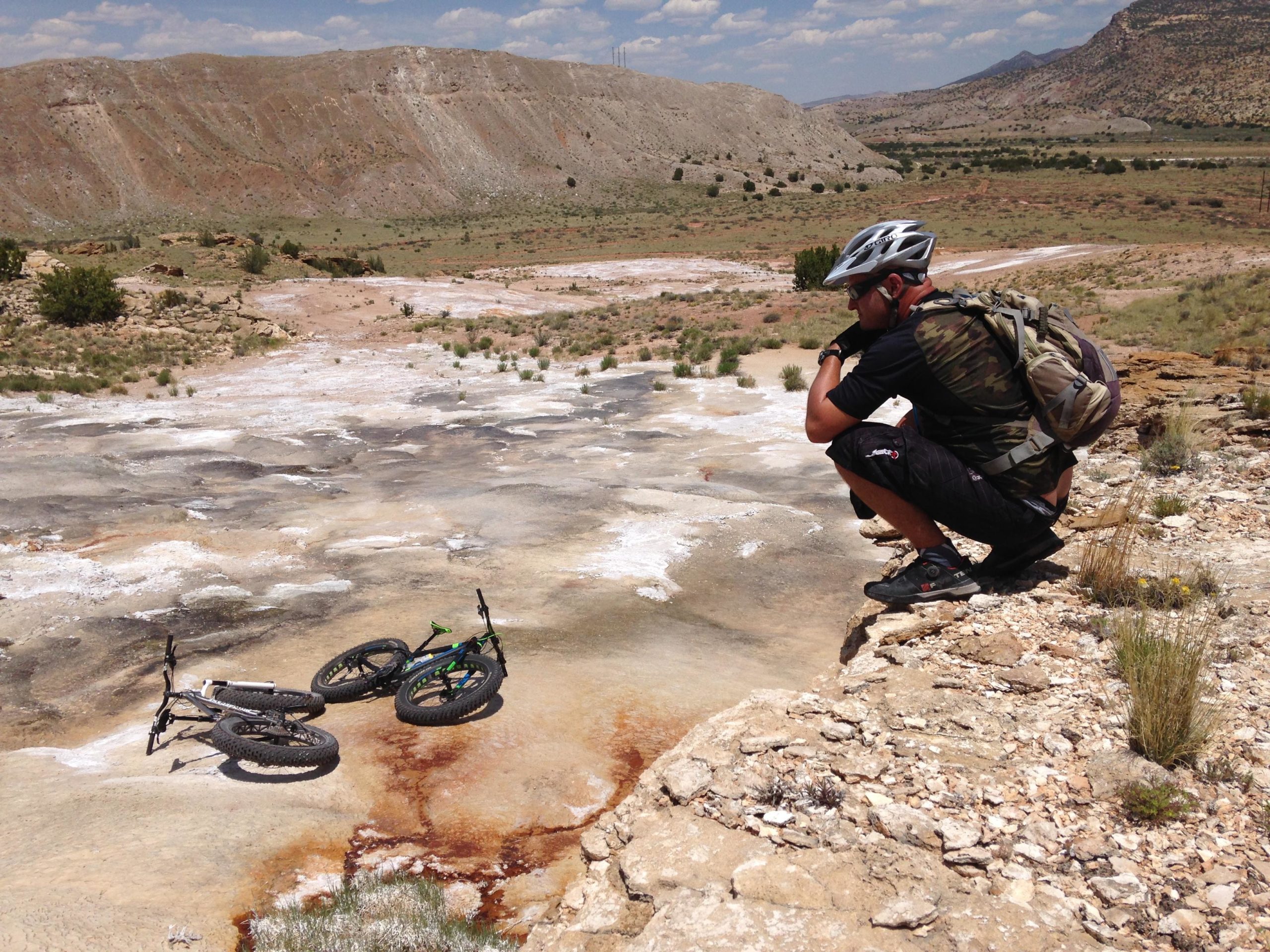 A man in a camouflage shirt and a helmet crouches on a rocky outcrop, looking towards two mountain bikes resting on a smooth, pale surface. The landscape around him features rocky terrain and sparse vegetation under a partly cloudy sky. White Ridge Bike Trails mountain bike trail.