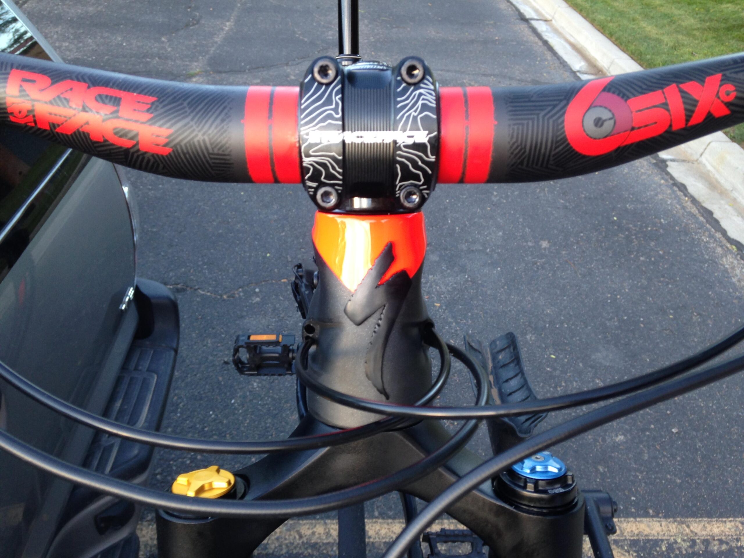 Specialized Stumpjumper FSR 6Fattie: Close-up view of a bicycle handlebar with a black and red color scheme, featuring brand logos. The handlebar is mounted on a black bicycle frame with visible cables and components. A grey vehicle is parked in the background, along with a patch of grass.