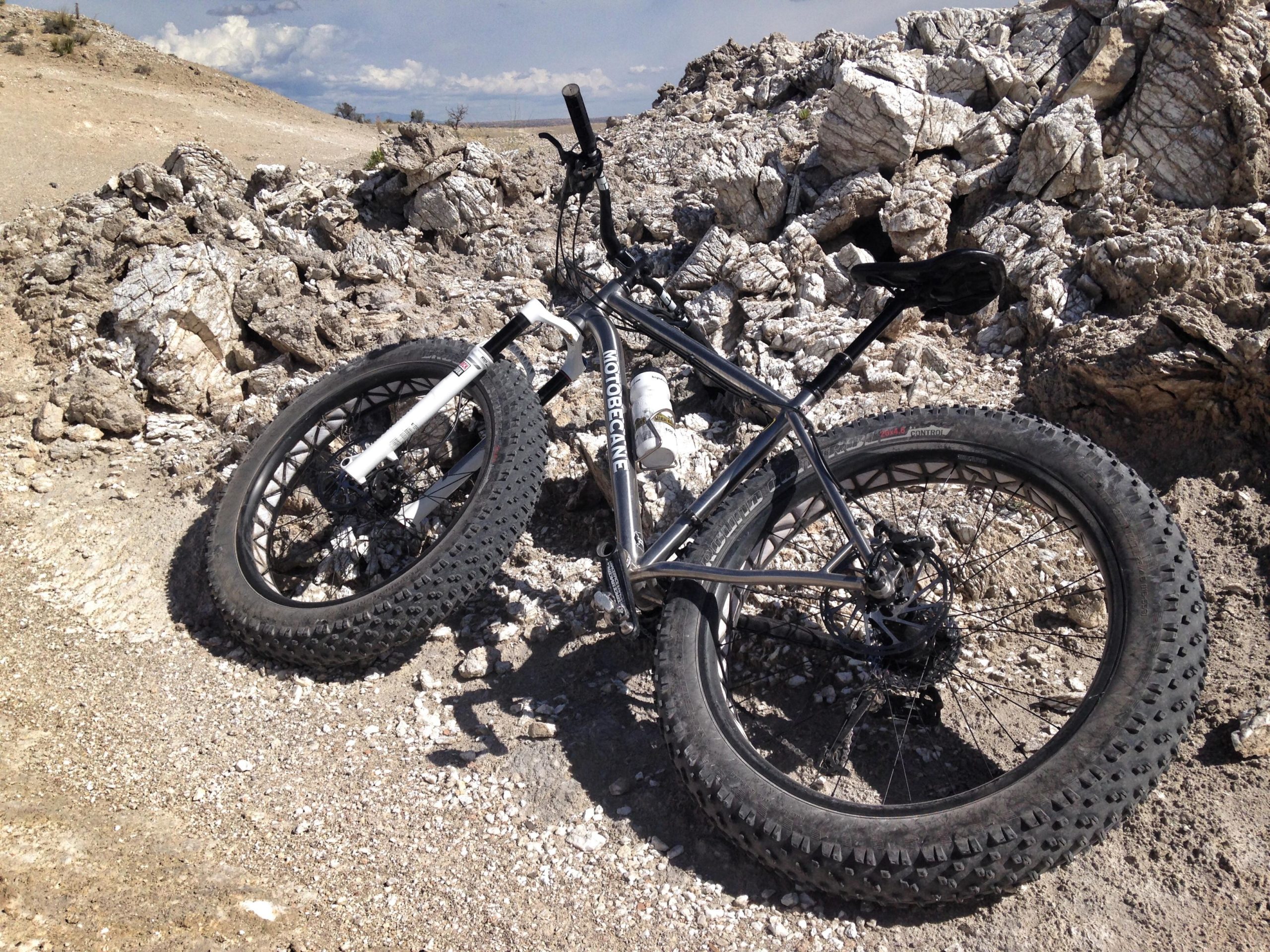 A black fat tire bike resting on rocky terrain under a partly cloudy sky. The bike is positioned on a gravel surface, surrounded by large stones and boulders. White Ridge Bike Trails mountain bike trail.