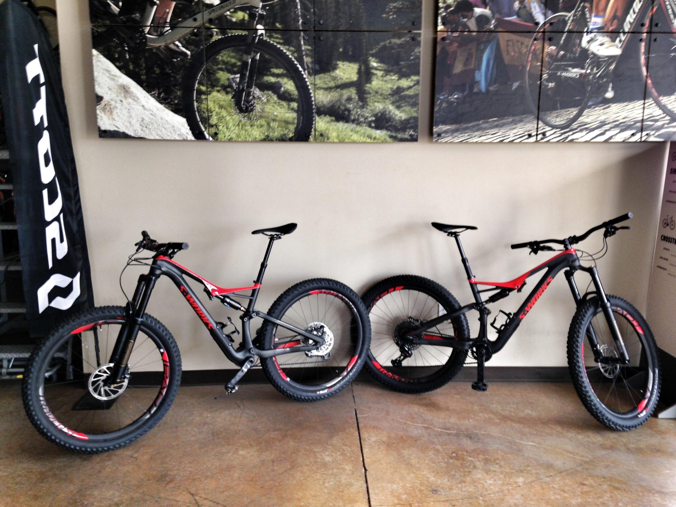 Two mountain bikes are displayed inside a shop. Both bikes have a sleek design with a black frame accented by red details. The bike on the left features a more robust front suspension, while the bike on the right has a slightly different frame design. Behind them, there are promotional images of biking activities on the wall, and a black flag with white lettering stands nearby. The floor is of polished concrete.