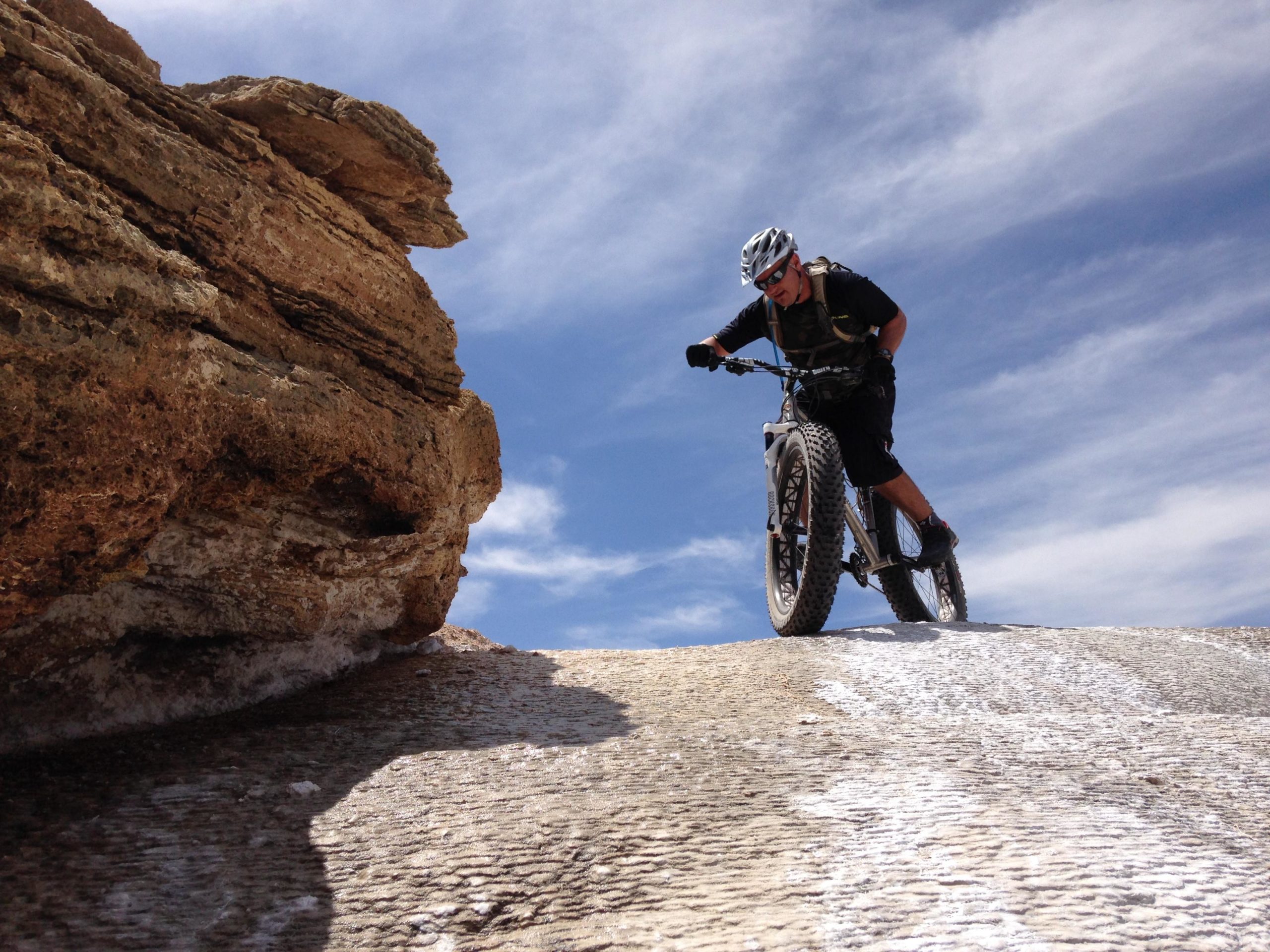 A mountain biker navigating a rocky terrain under a blue sky with white clouds. The biker is positioned on a large rock formation, displaying a dynamic posture while riding a fat bike. White Ridge Bike Trails mountain bike trail.