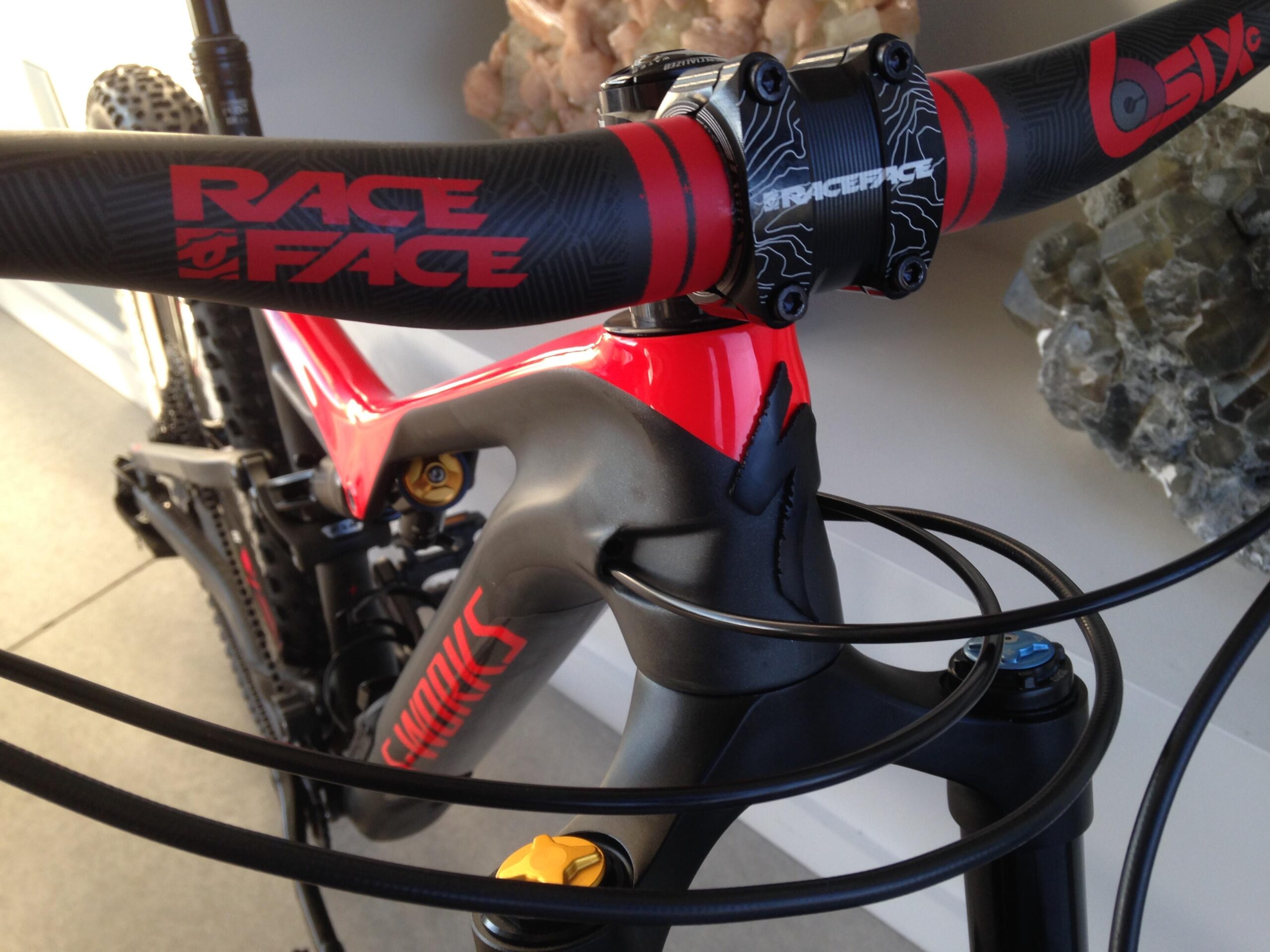 Specialized Stumpjumper FSR 6Fattie: Close-up view of a red and black mountain bike, featuring a prominent "Race Face" handlebar and stem. The frame has a sleek design with "S-Works" branding, and visible components include hydraulic cables and a yellow adjustment knob. The background shows a minimalistic indoor setting with decorative stones.