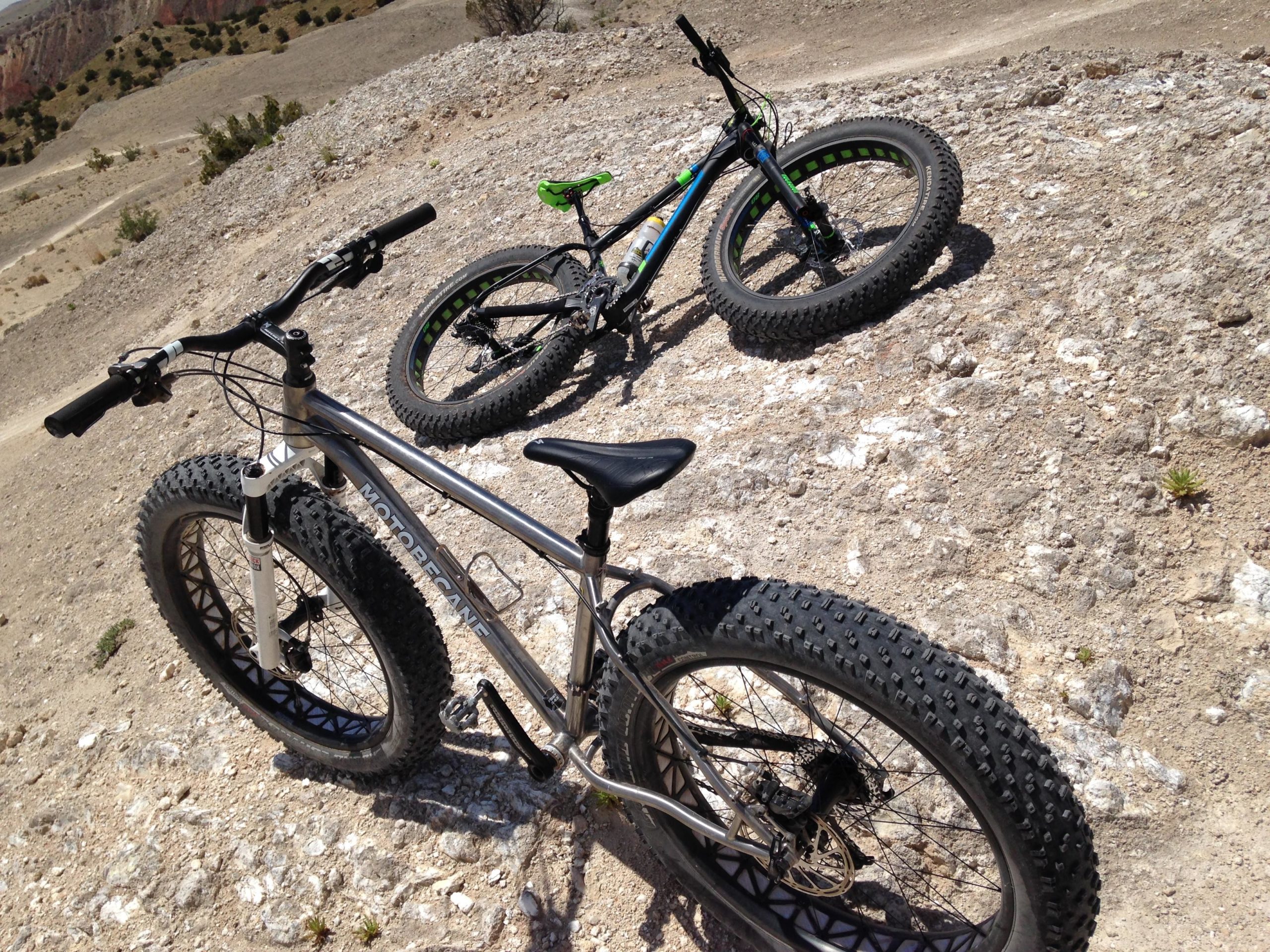 Two bicycles are parked on a rocky, barren terrain under bright sunlight. One bike has a silver frame and wide, textured tires, while the other is multicolored with a black frame and green accents. The background features a hilly landscape, suggesting a rugged outdoor cycling environment. White Ridge Bike Trails mountain bike trail.