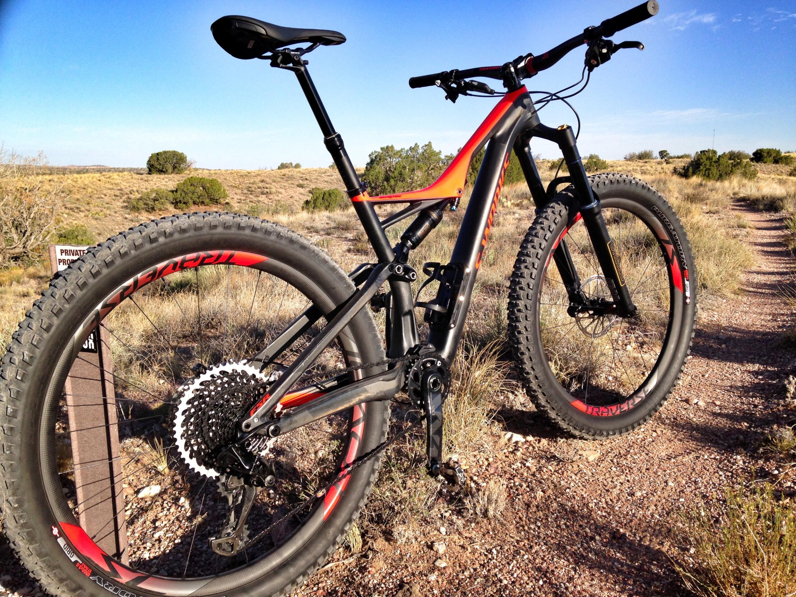 Specialized Stumpjumper FSR 6Fattie: A mountain bike with a sleek design and wide tires is positioned on a dirt trail surrounded by natural terrain. The bike features a black and orange color scheme, with prominent branding and a scenic background of shrubs and distant hills under a clear blue sky. A sign indicating "Private Property" is visible in the foreground.