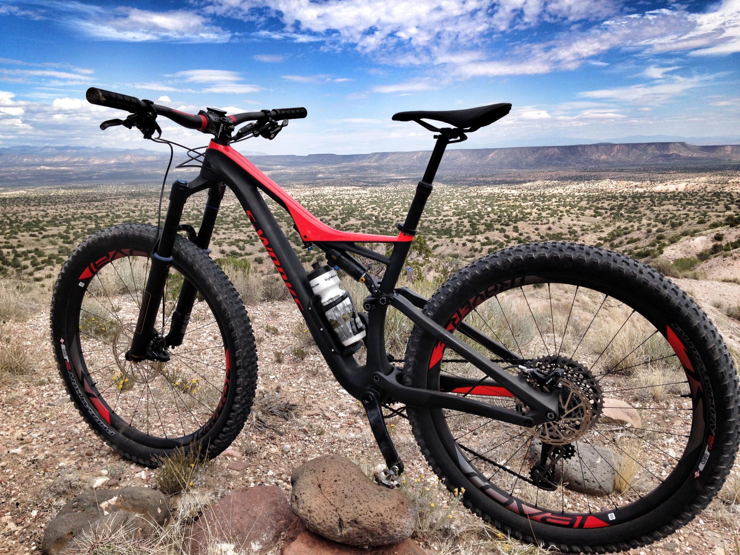 A mountain bike with a sleek black and red design is parked on rocky terrain, overlooking a vast landscape of rolling hills and sparse vegetation under a bright blue sky with white clouds. Mariposa Fat Bike Trails mountain bike trail.