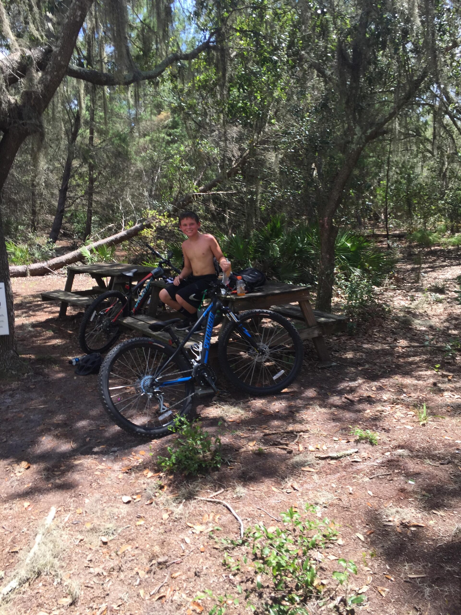Specialized Rockhopper 29: A young boy sitting on a mountain bike in a wooded area, enjoying a sunny day. He is shirtless and smiling, with a picnic table visible in the background surrounded by trees and greenery.