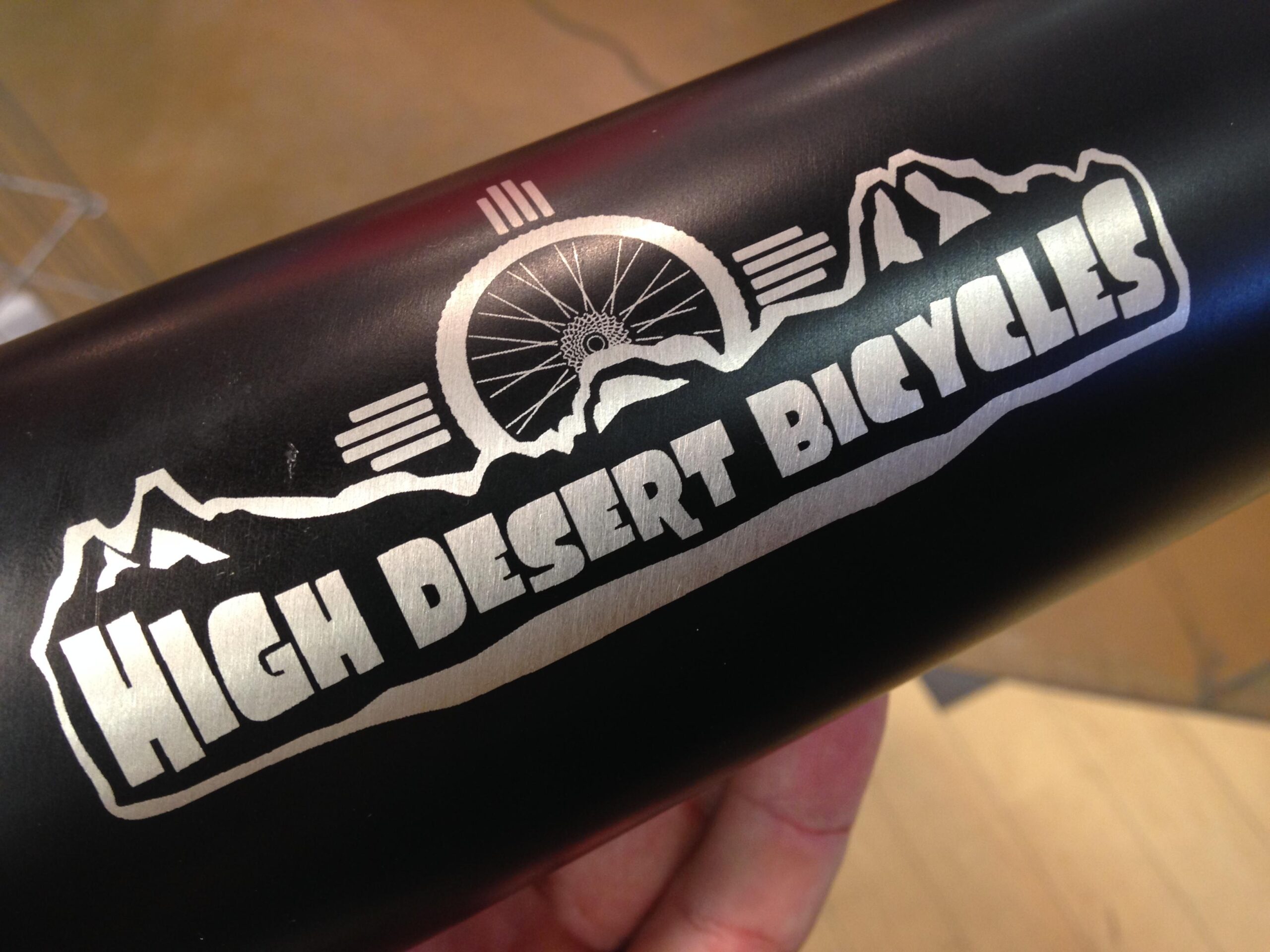 A close-up image of a black metal bike frame featuring the engraved logo of "High Desert Bicycles." The logo includes stylized text and a design with mountains and a bicycle wheel. The background shows a blurred interior space, suggesting a bicycle shop or retail environment.