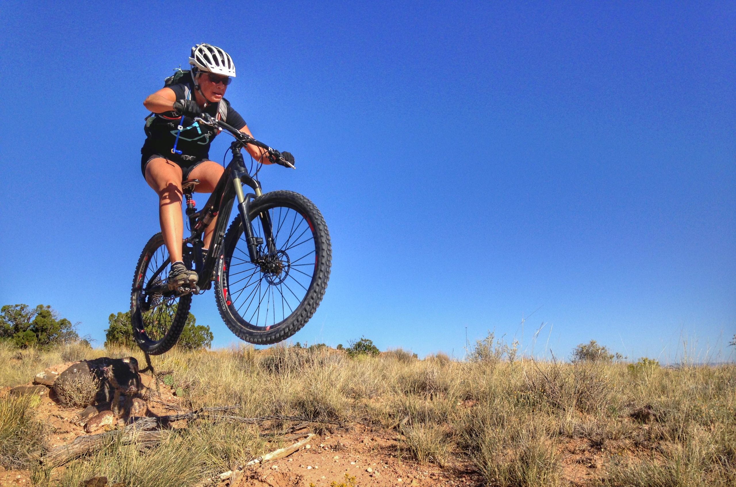 A mountain biker capturing air while performing a jump on a trail, surrounded by dry grass and shrubs under a clear blue sky. The cyclist is wearing a helmet and riding a black mountain bike with large tires. Mariposa Fat Bike Trails mountain bike trail.