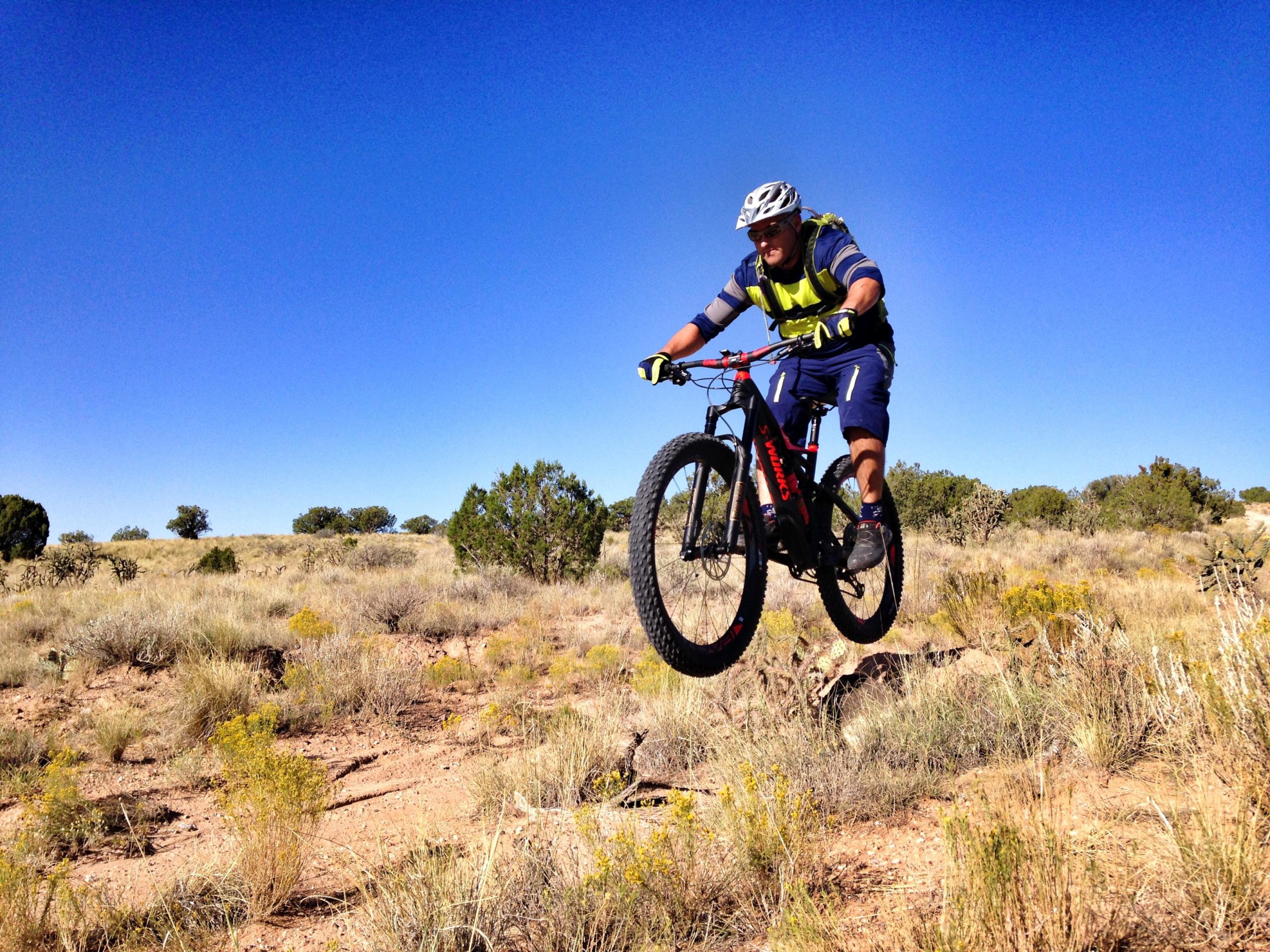 A mountain biker in mid-air, performing a jump over a rocky terrain, surrounded by dry grass and sparse shrubs under a clear blue sky. The cyclist is wearing a helmet, a reflective vest, and protective gear, showcasing an adventurous outdoor activity in a natural landscape. Mariposa Fat Bike Trails mountain bike trail.