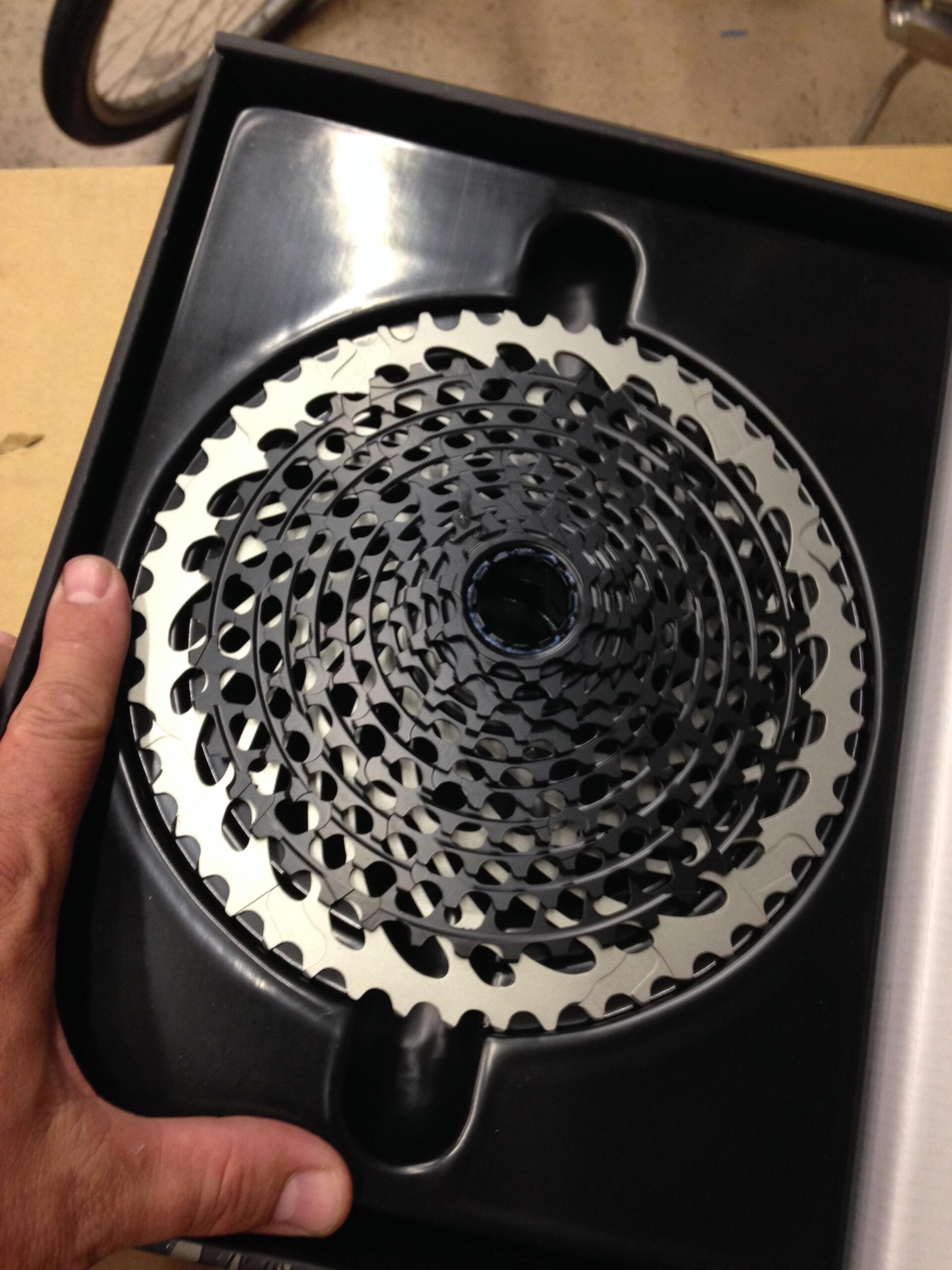 A close-up image of a hand holding a bicycle cassette, which is placed inside a black plastic packaging. The cassette consists of interlocking gears with a mixture of black and silver colors, and the background shows a hint of a bicycle wheel.