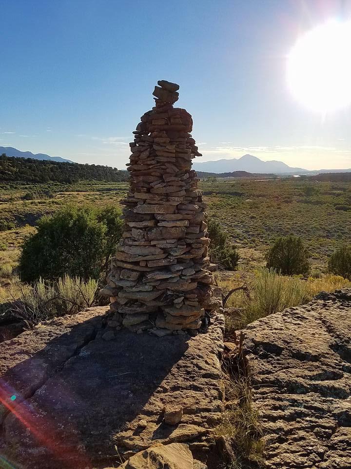 A tall stone cairn standing on a rocky outcrop, with a scenic view of rolling hills and mountains in the background. The sun is low in the sky, casting warm light across the landscape and creating lens flares. Green vegetation is visible in the foreground, contrasting with the earthy tones of the rocks. Phil's World mountain bike trail.