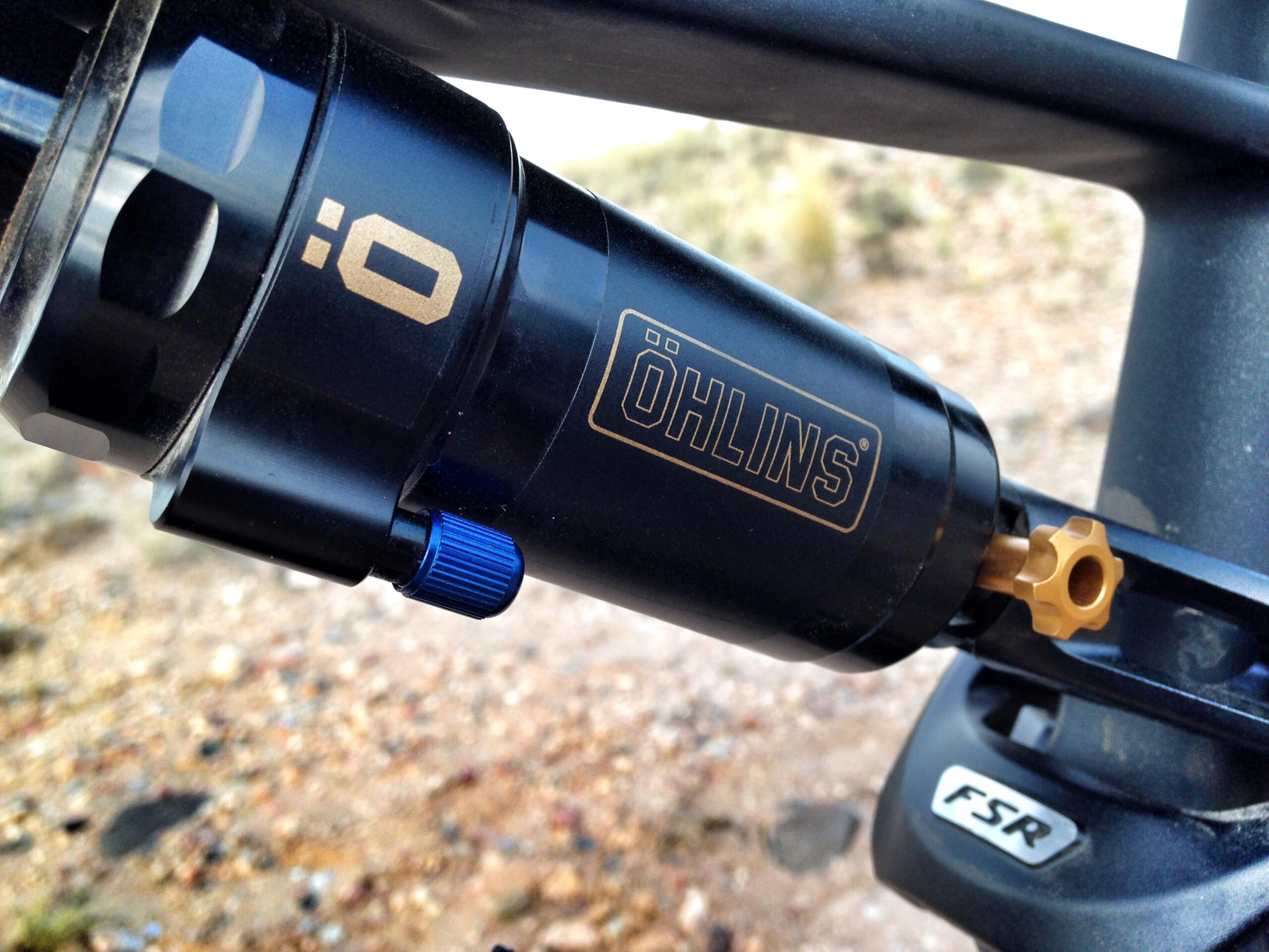 Specialized Stumpjumper FSR 6Fattie: A close-up image of a black Öhlins shock absorber with a gold logo, featuring a blue adjustment knob and part of a mountain bike frame with a visible FSR logo in the background. The setting appears rugged and outdoorsy, suggesting a focus on mountain biking components.