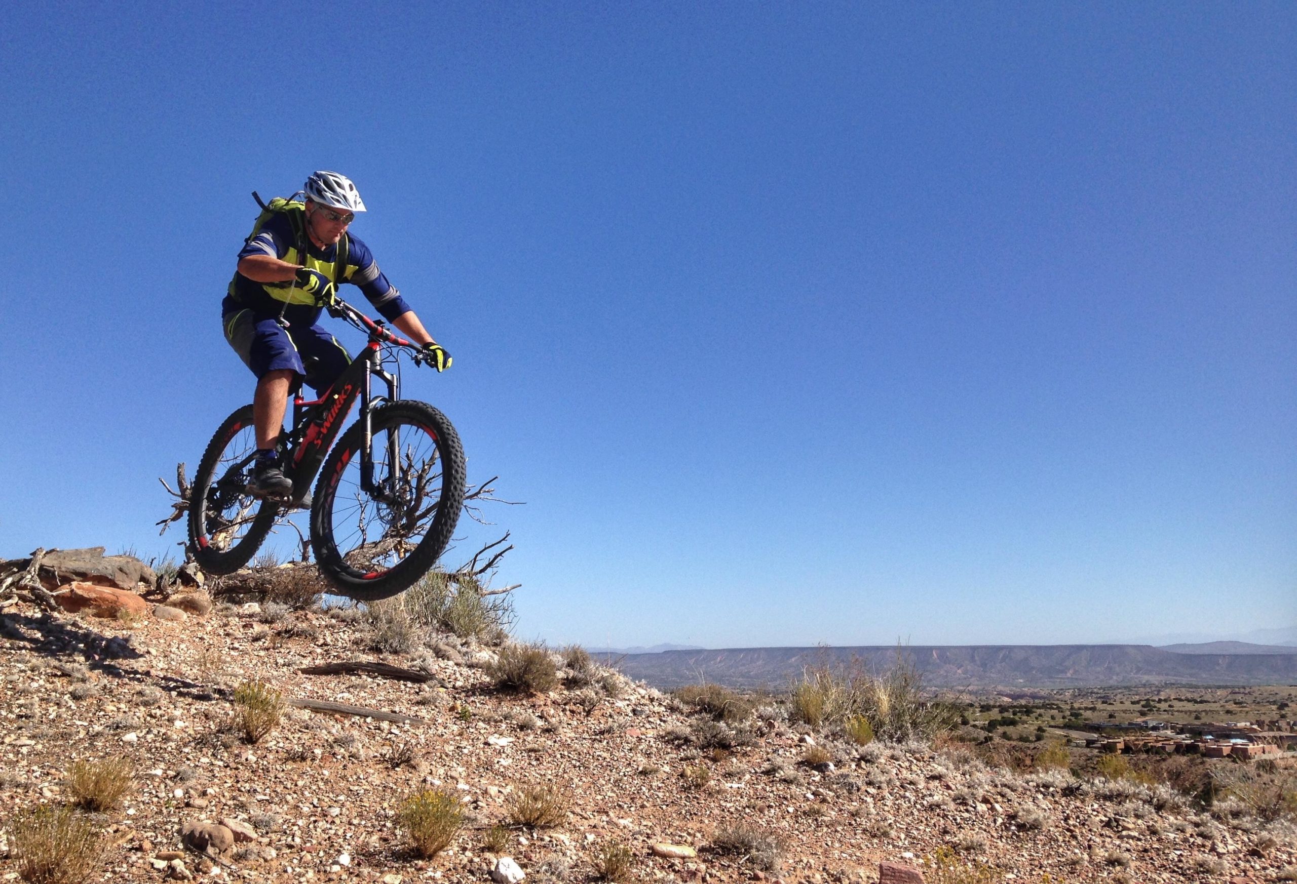 A mountain biker in protective gear jumps off a rocky terrain, demonstrating excitement and skill against a clear blue sky and distant mountainous landscape. Mariposa Fat Bike Trails mountain bike trail.