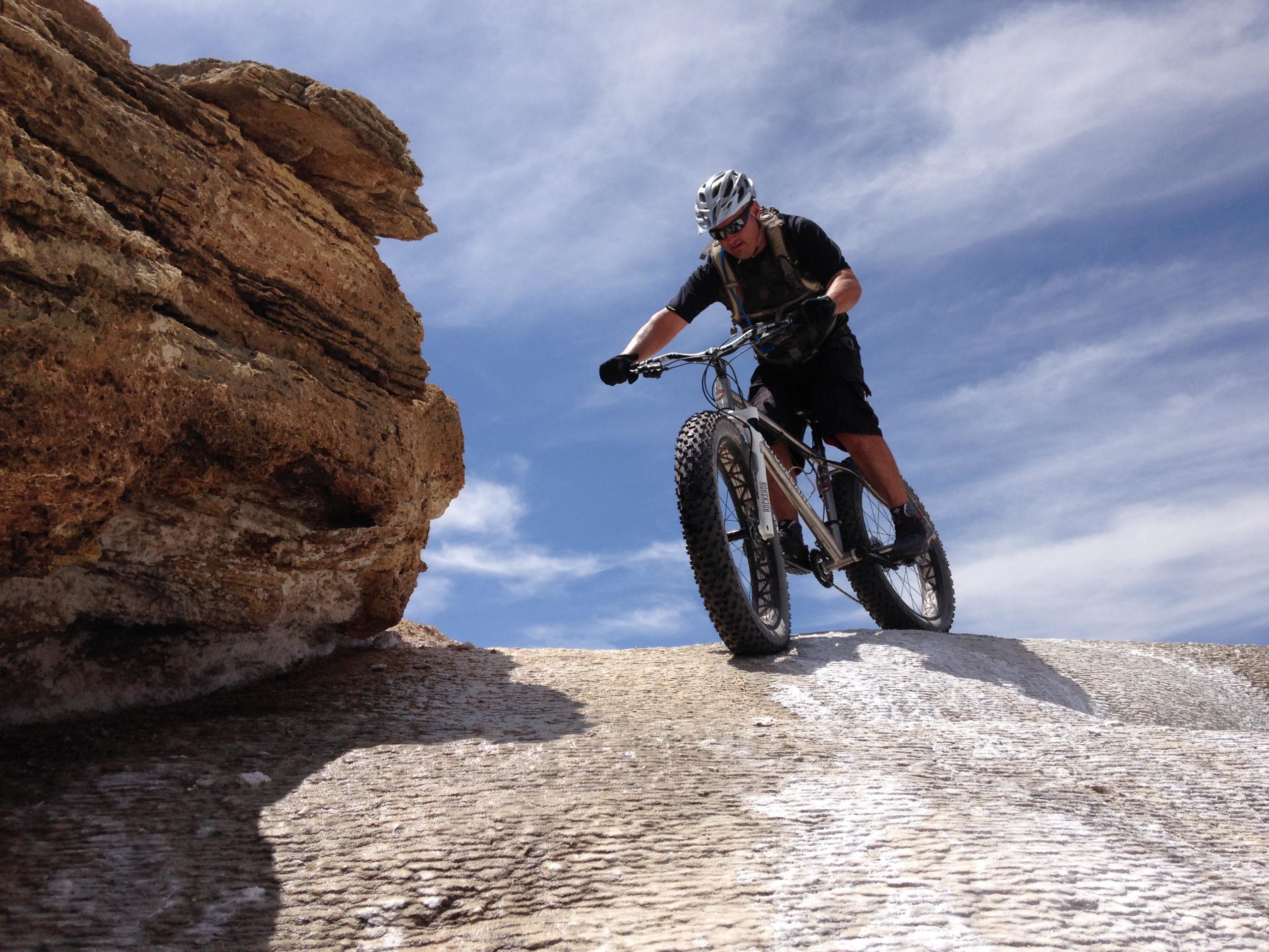 A mountain biker rides a fat bike on a rocky terrain under a blue sky with wispy clouds. The biker is leaning forward, focusing on navigating the challenging surface, while a large rock formation is visible on the left side of the image. White Ridge Bike Trails mountain bike trail.