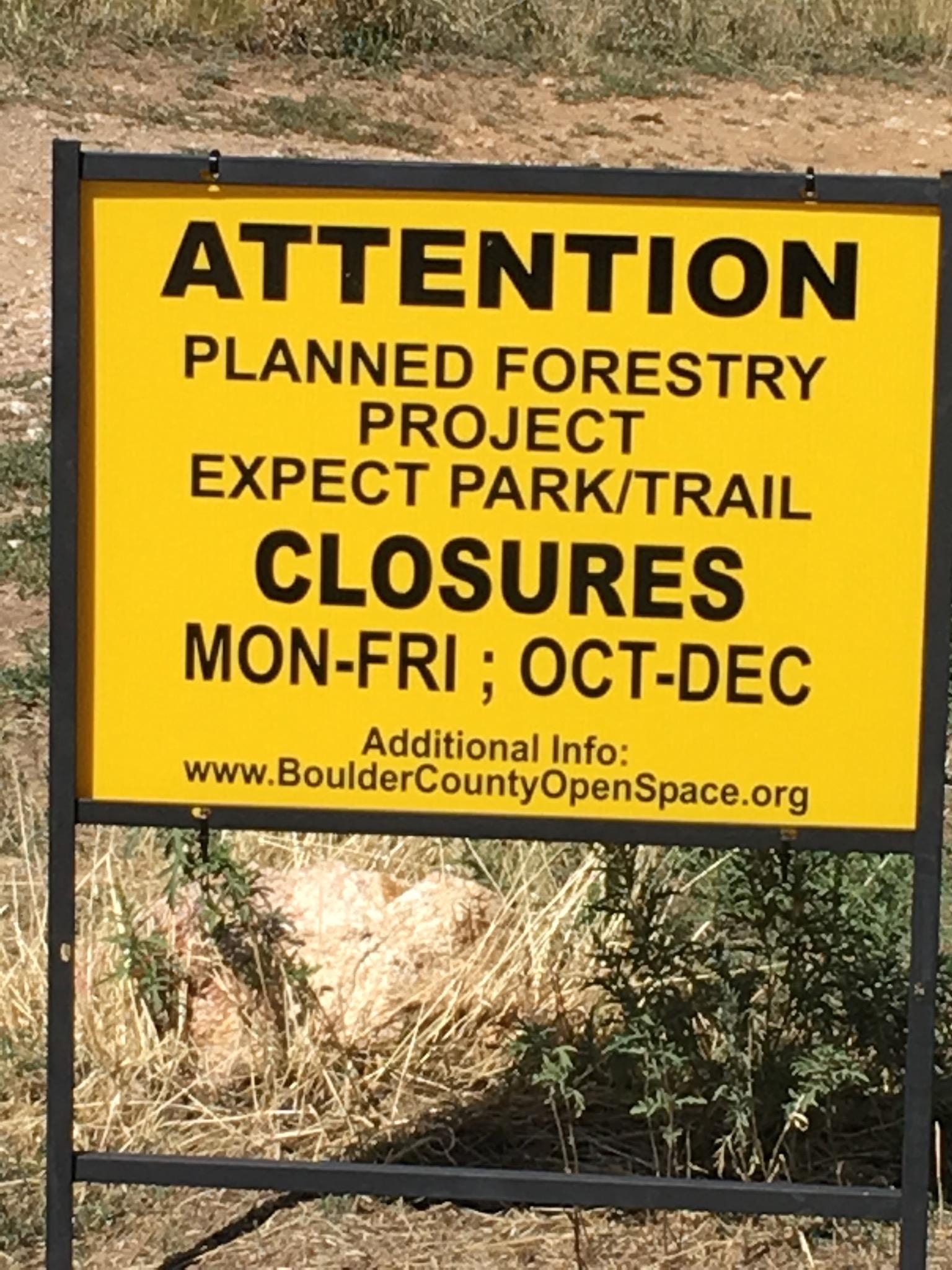 A bright yellow sign indicating a planned forestry project with warnings about park and trail closures from Monday to Friday during the months of October to December. The sign includes a web address for additional information. Betasso Preserve mountain bike trail.