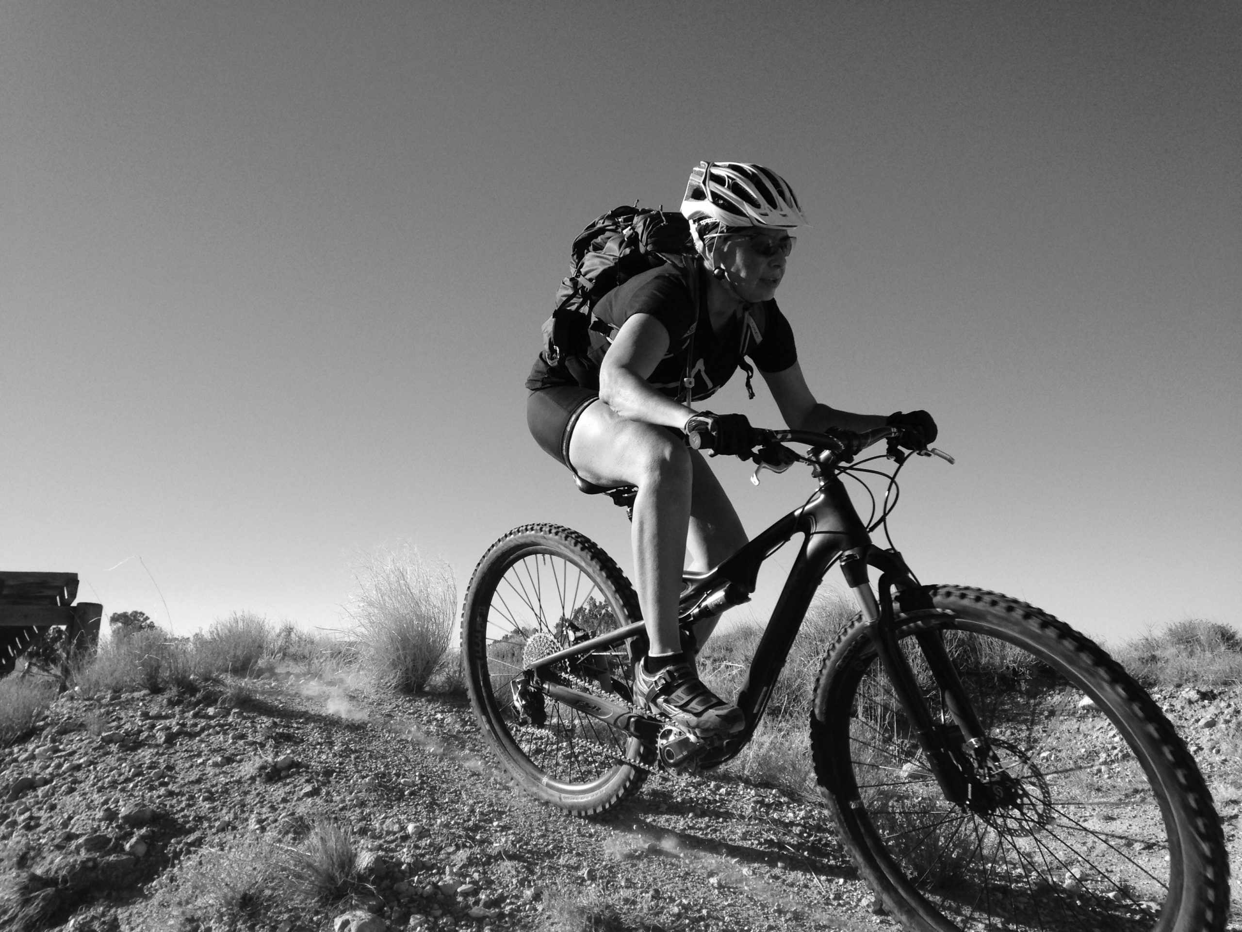 A cyclist riding a mountain bike downhill on a rocky terrain, wearing a helmet and sunglasses, with a backpack. The image is in black and white, showcasing the motion and dust kicked up by the bike. Sparse vegetation and a clear sky are in the background. Mariposa Fat Bike Trails mountain bike trail.