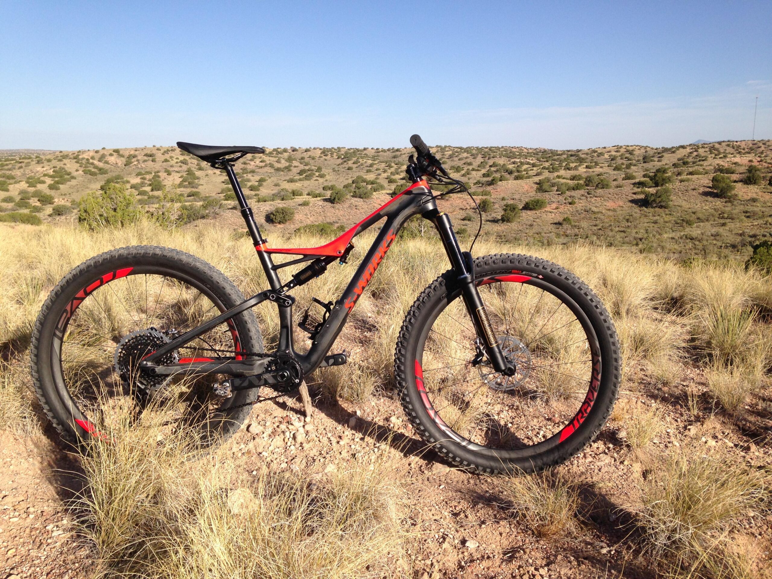 Specialized Stumpjumper FSR 6Fattie: A black and red mountain bike is parked on a sandy trail surrounded by tall grass. In the background, rolling hills and sparse vegetation extend under a clear blue sky. The bike features wide tires and a sleek frame, showcasing its suitability for off-road riding.