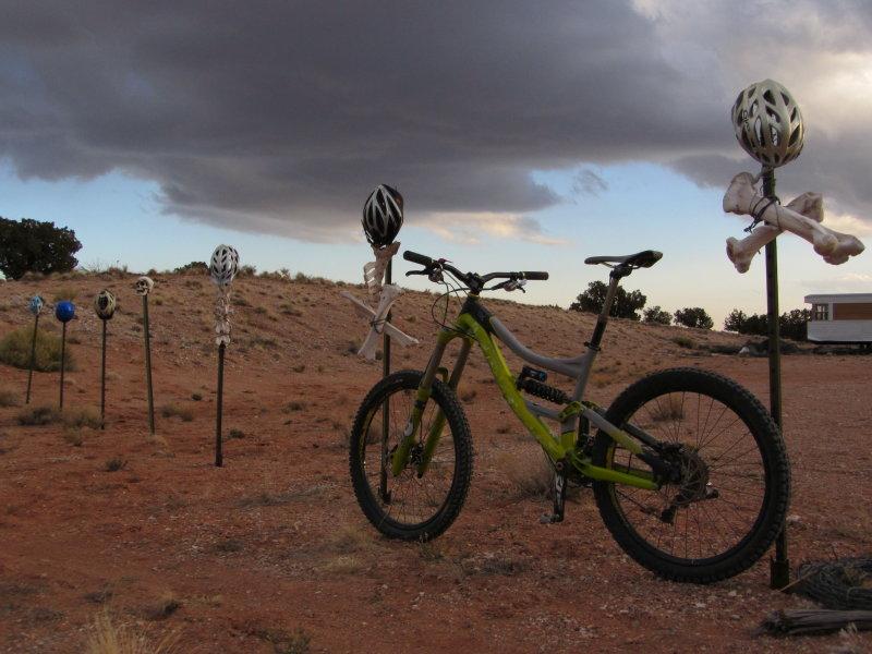 A green mountain bike rests on sandy terrain, alongside a row of poles topped with various bicycle helmets, some attached to skeletal figures. The sky is overcast with dark clouds, creating a dramatic backdrop. In the distance, a trailer is partially visible among sparse vegetation. Mariposa Fat Bike Trails mountain bike trail.