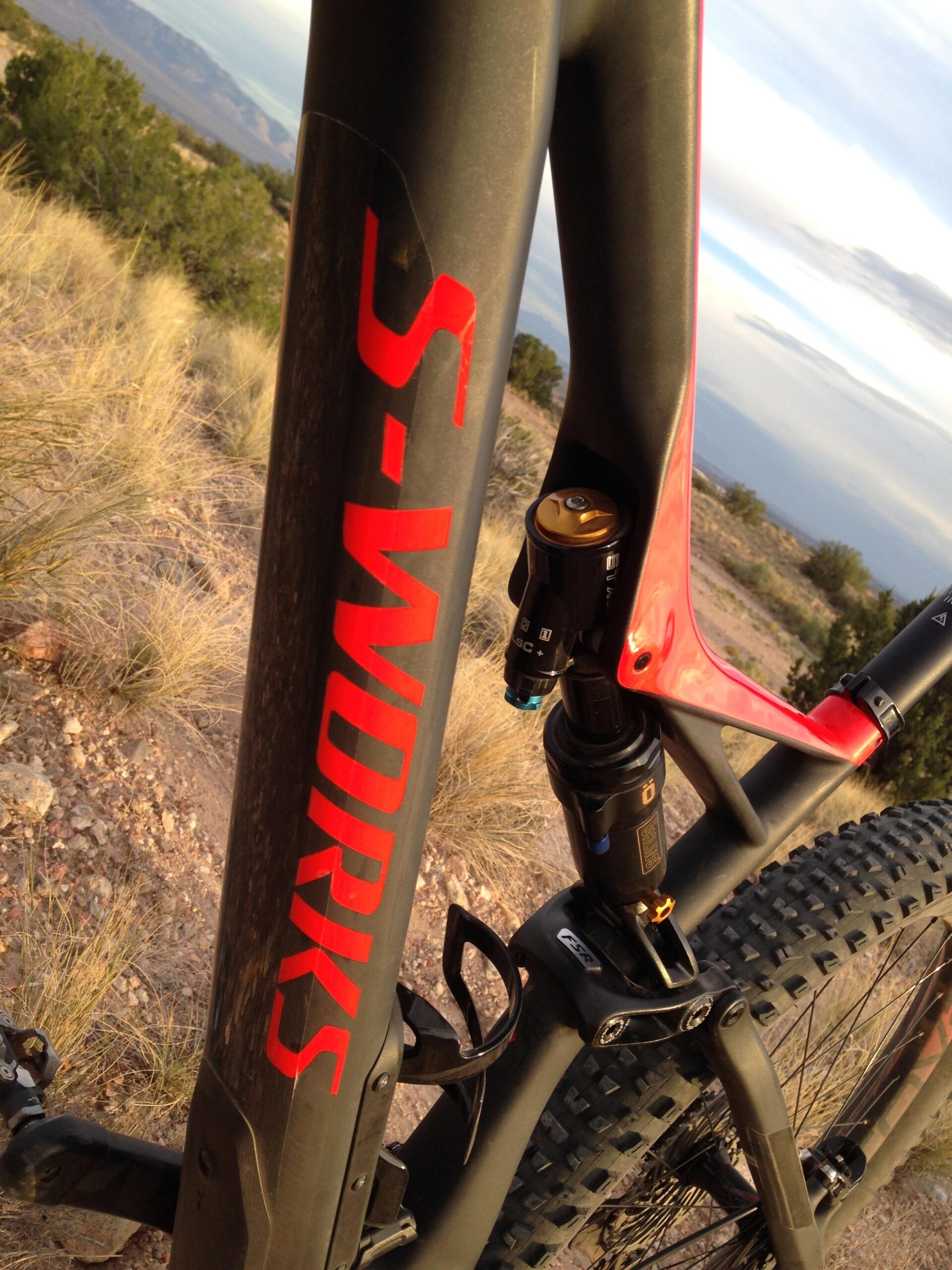 Specialized Stumpjumper FSR 6Fattie: Close-up of a mountain bike frame featuring the "S-Works" logo in bold red lettering, showcasing the suspension system and rear shock. The background includes a natural landscape with grass and distant mountains under a cloudy sky.