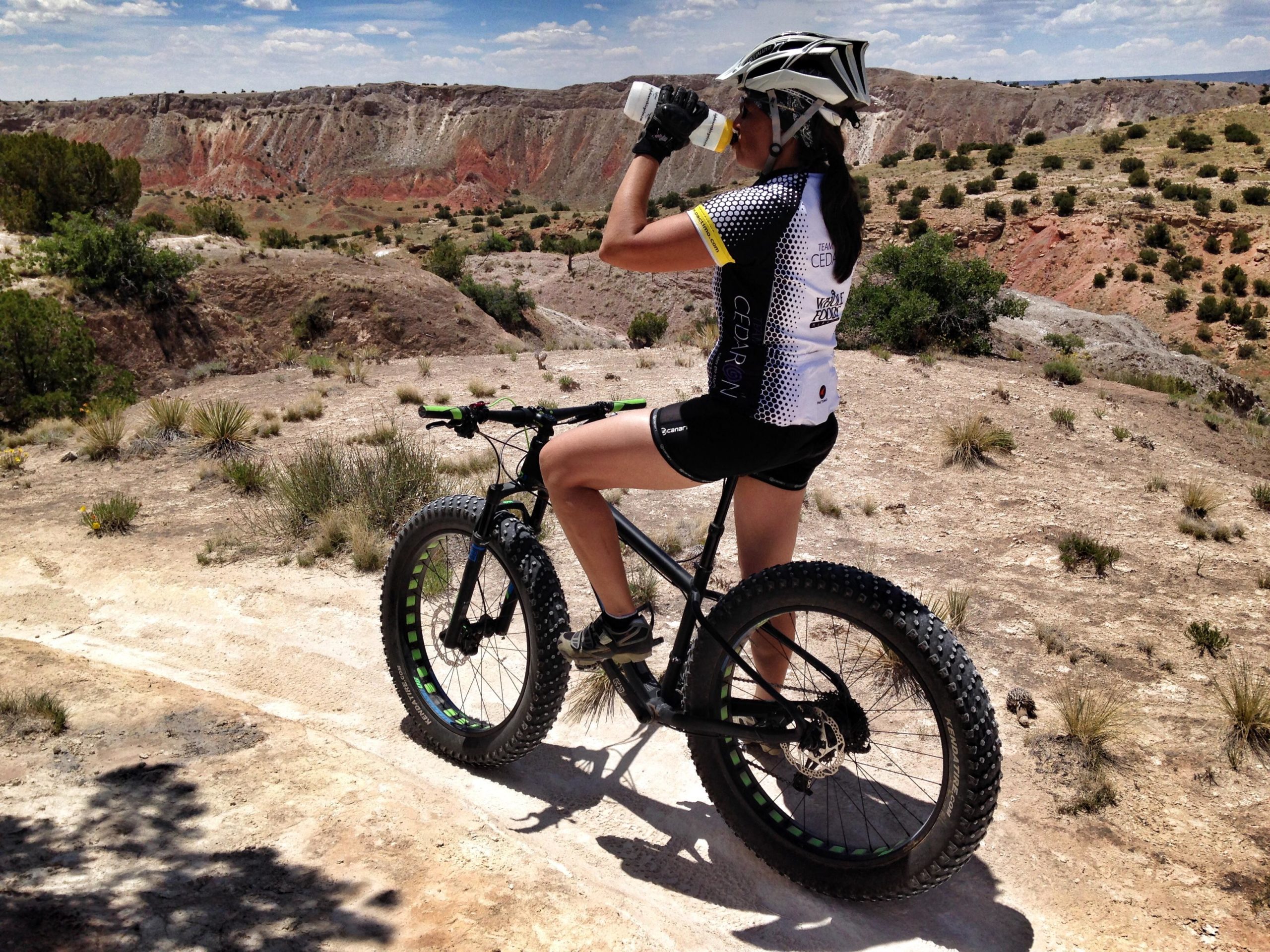 A person wearing a cycling jersey and helmet stands beside a fat tire mountain bike on a rocky trail, taking a drink from a water bottle. The landscape features rolling hills with sparse vegetation and colorful rock formations in the background under a blue sky with scattered clouds. White Ridge Bike Trails mountain bike trail.