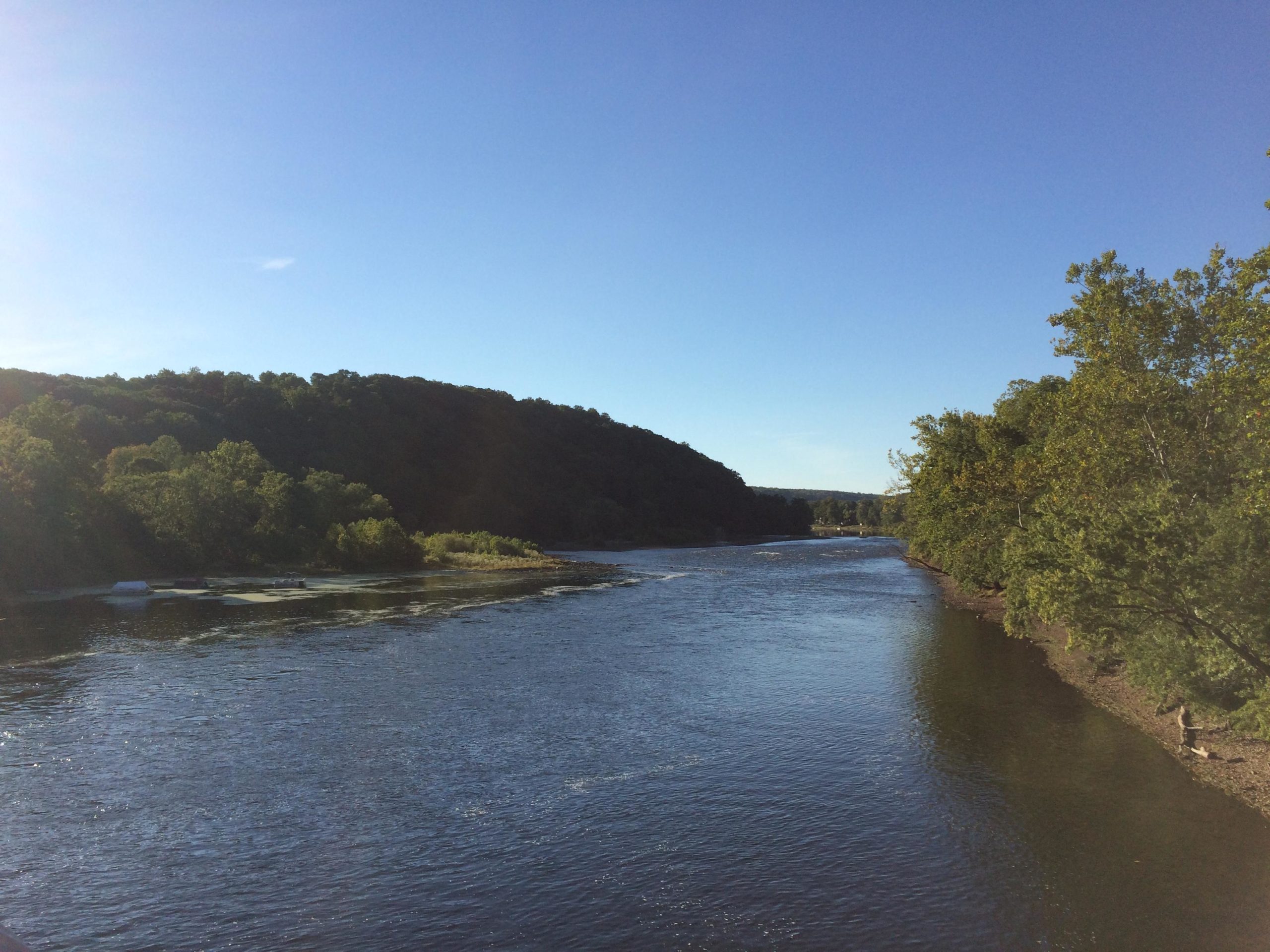 A serene view of a river winding through a landscape of lush green trees and hills under a clear blue sky. The water reflects the sunlight, adding to the tranquil ambiance of the scene. Bridge To Bridge - D&r Canal mountain bike trail.