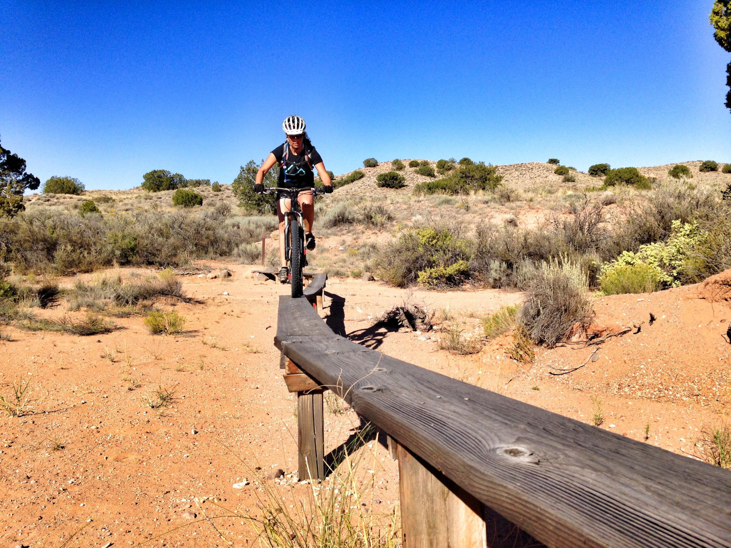 A mountain biker skillfully rides along a narrow wooden plank trail in a desert landscape under a clear blue sky. Surrounding the trail are patches of shrubs and small trees, with sandy terrain visible in the foreground. The biker is wearing a helmet and gear, demonstrating an active outdoor lifestyle. Mariposa Fat Bike Trails mountain bike trail.