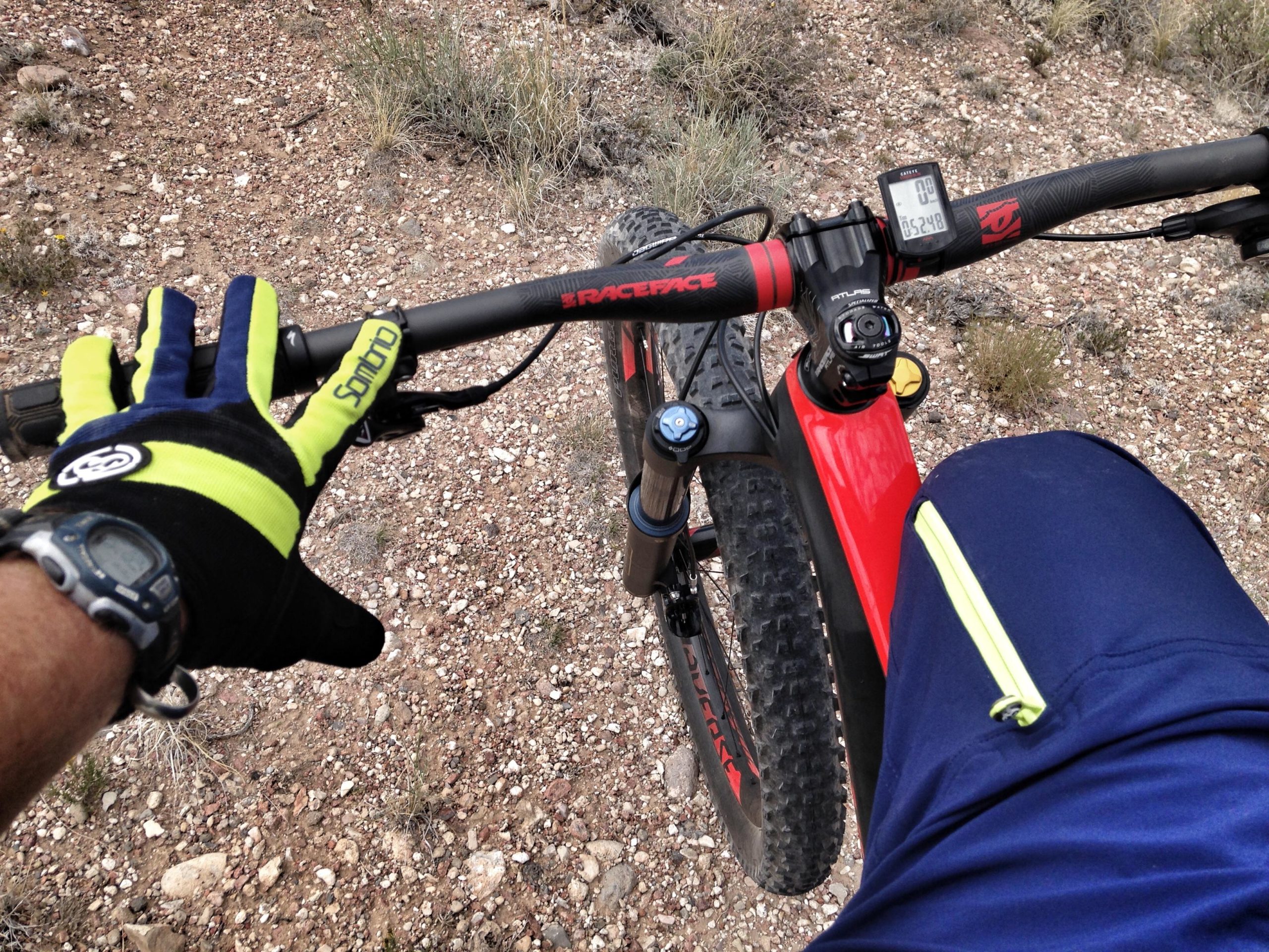 Person riding a mountain bike, focusing on the handlebars with one gloved hand resting on the grips. The bike features a digital display showing the time or speed, and the rider is wearing a watch and sportswear. The setting is rocky and natural, indicative of a biking trail. Mariposa Fat Bike Trails mountain bike trail.