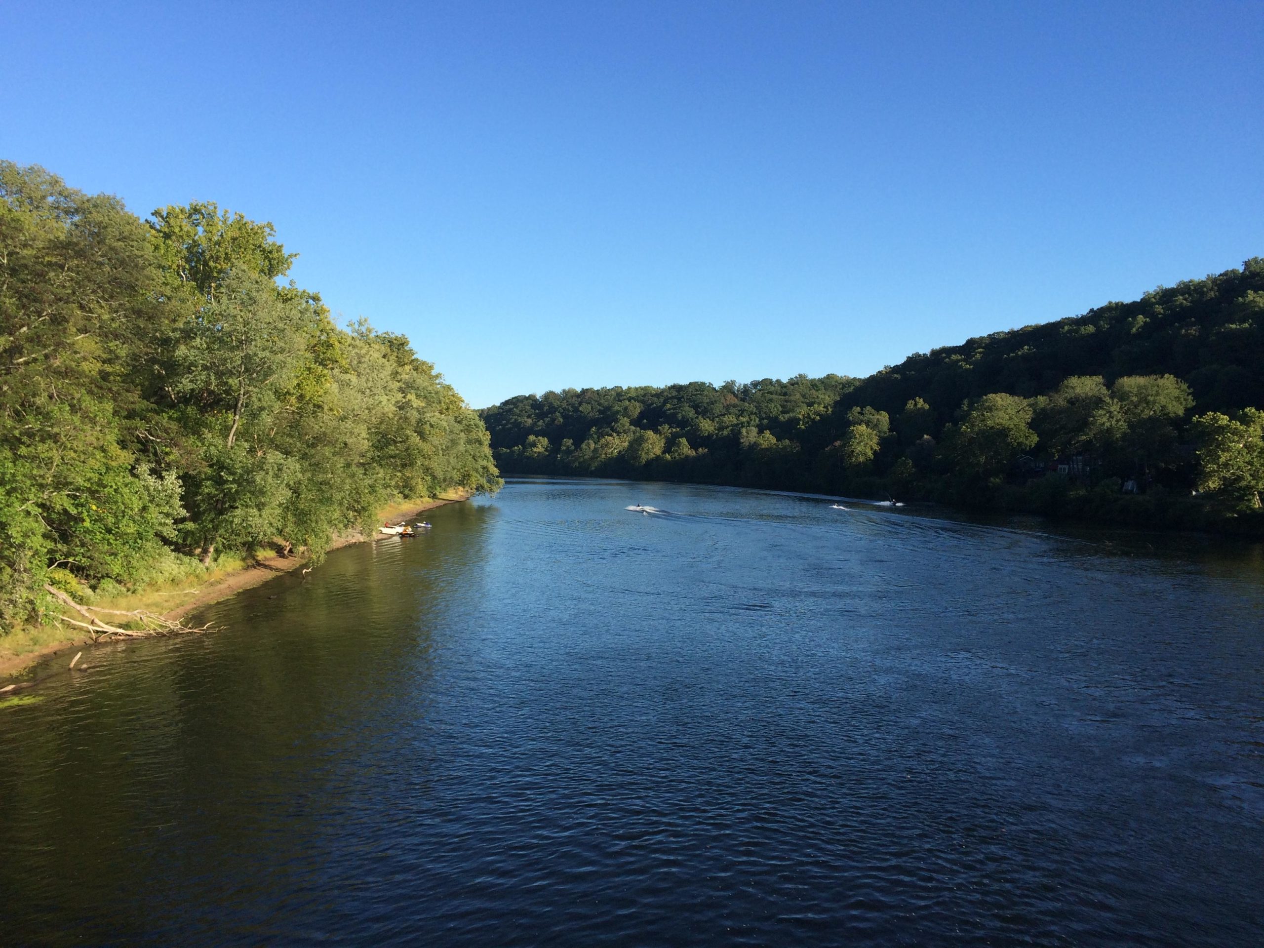 A serene river landscape under a clear blue sky, with lush greenery lining the banks. Small boats can be seen on the water, creating gentle ripples as they navigate the river. The scene captures the tranquil beauty of nature and the peaceful ambiance of a sunny day. Bridge To Bridge - D&r Canal mountain bike trail.